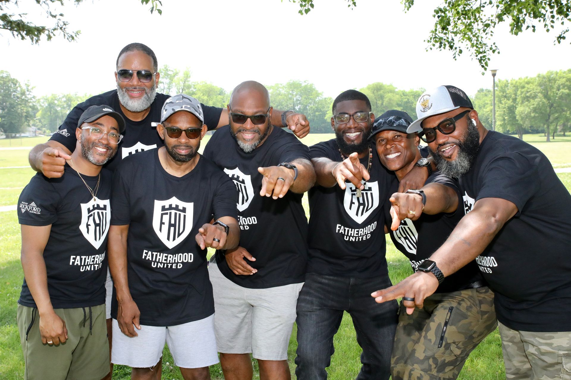 Group of seven people wearing black shirts and hats outdoors, pointing towards the camera.