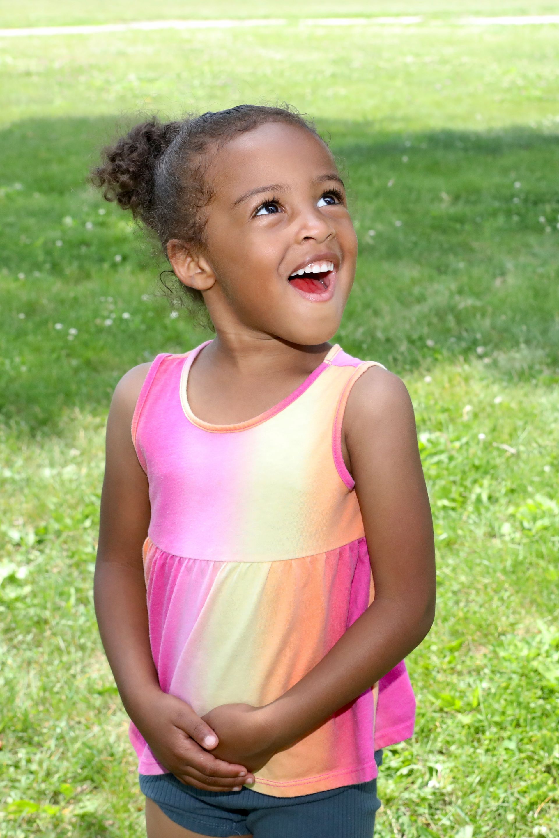 Young child with curly hair and a colorful shirt, laughing and looking upward in a grassy area.