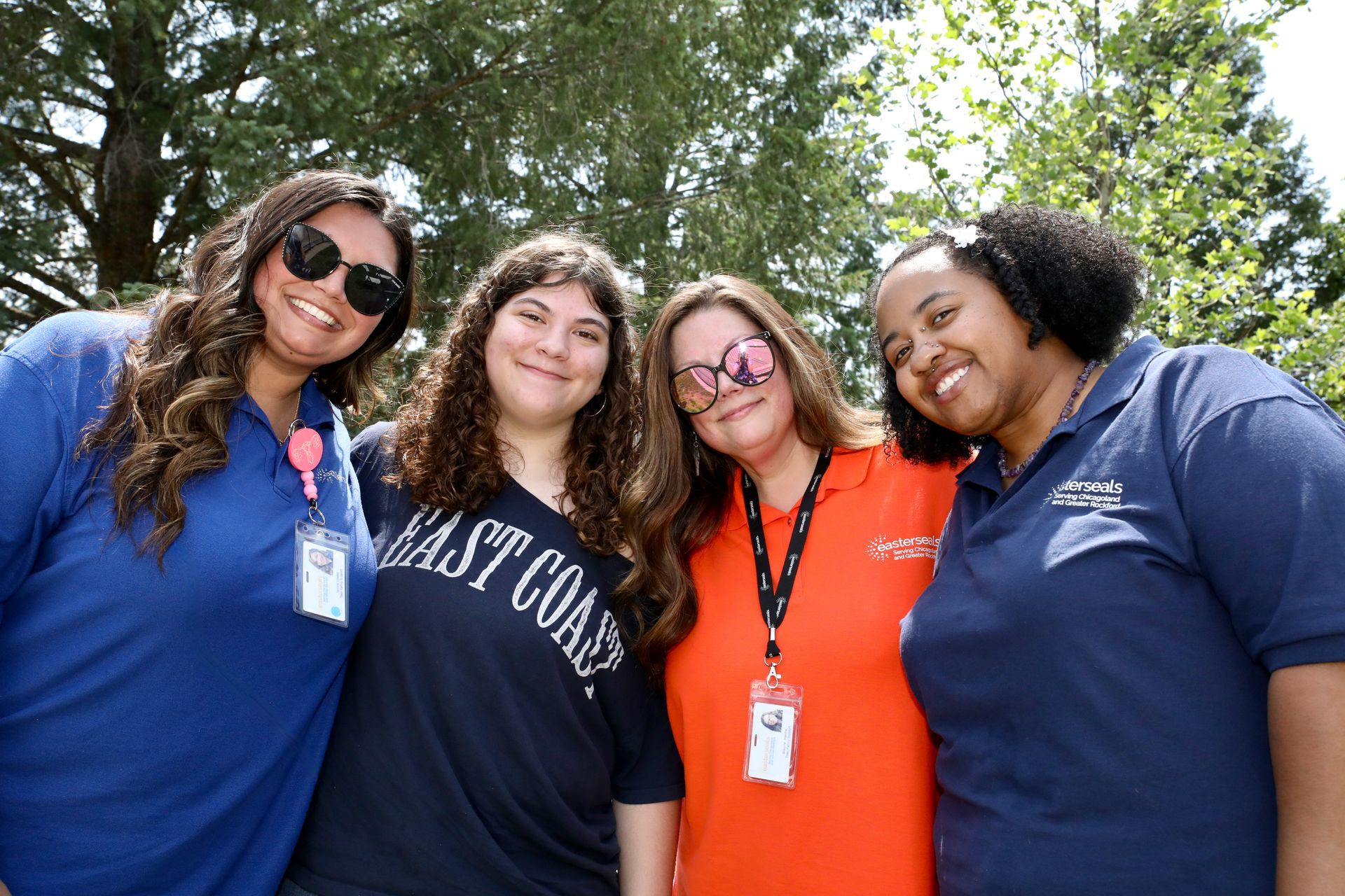 Four people smiling and posing outdoors; two wear blue shirts, one orange, all with lanyards.