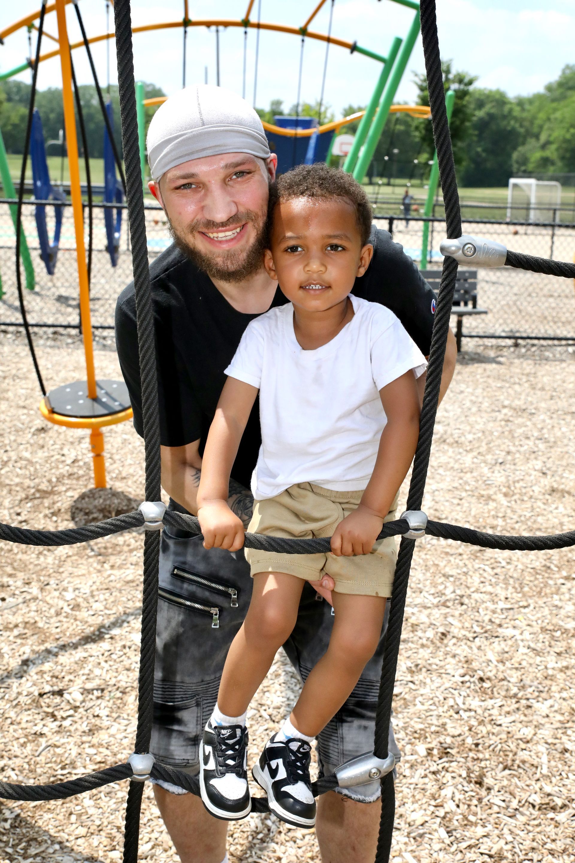 Man and child on playground climbing structure, smiling. Child wears white shirt, khaki shorts, black and white shoes.