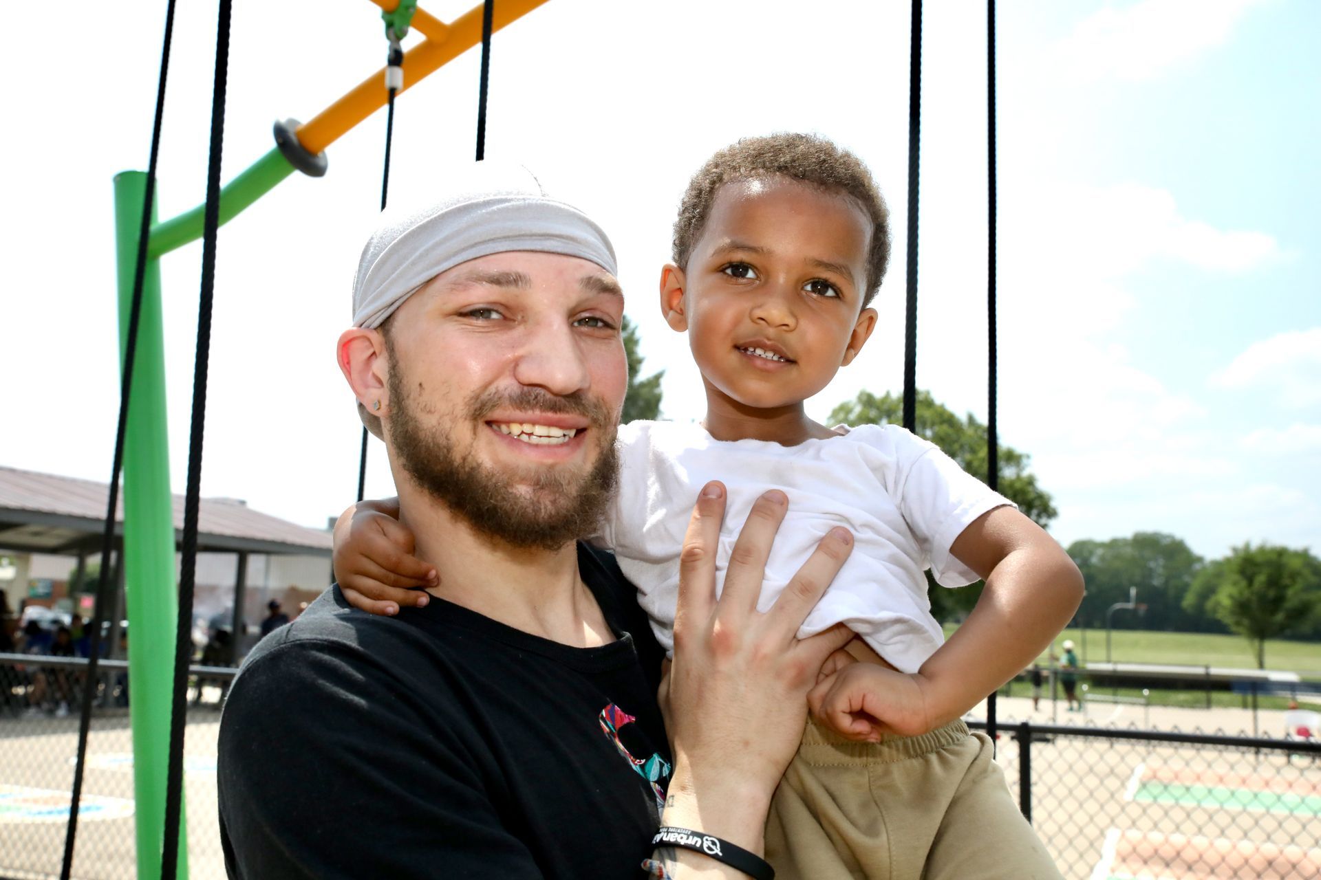Man holding child smiling at a playground. Green, yellow equipment. Child is wearing a white shirt and tan pants.