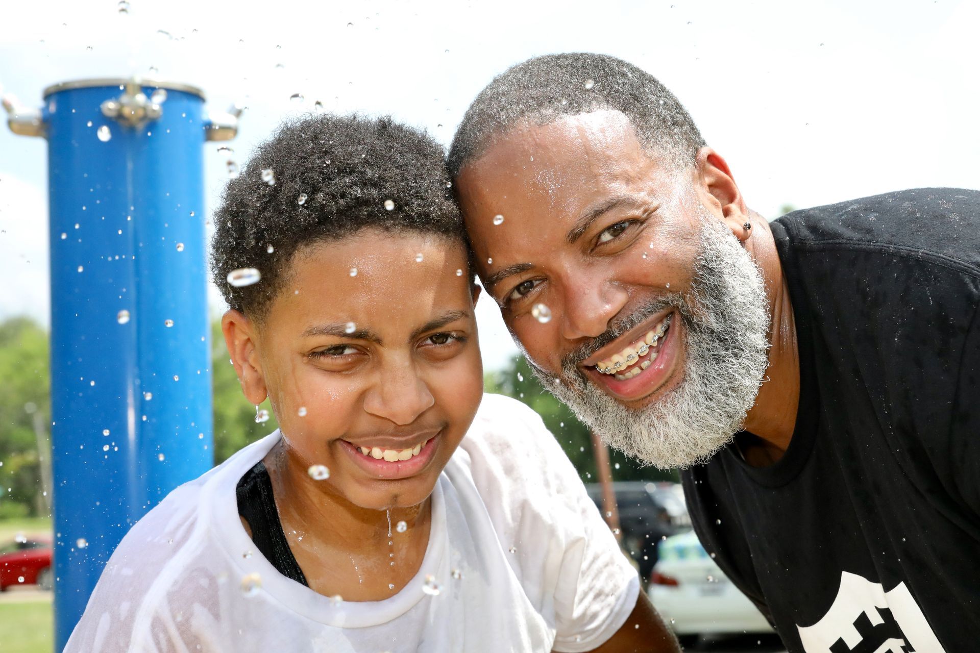 Father and child getting wet by a water fountain outdoors, smiling at the camera.