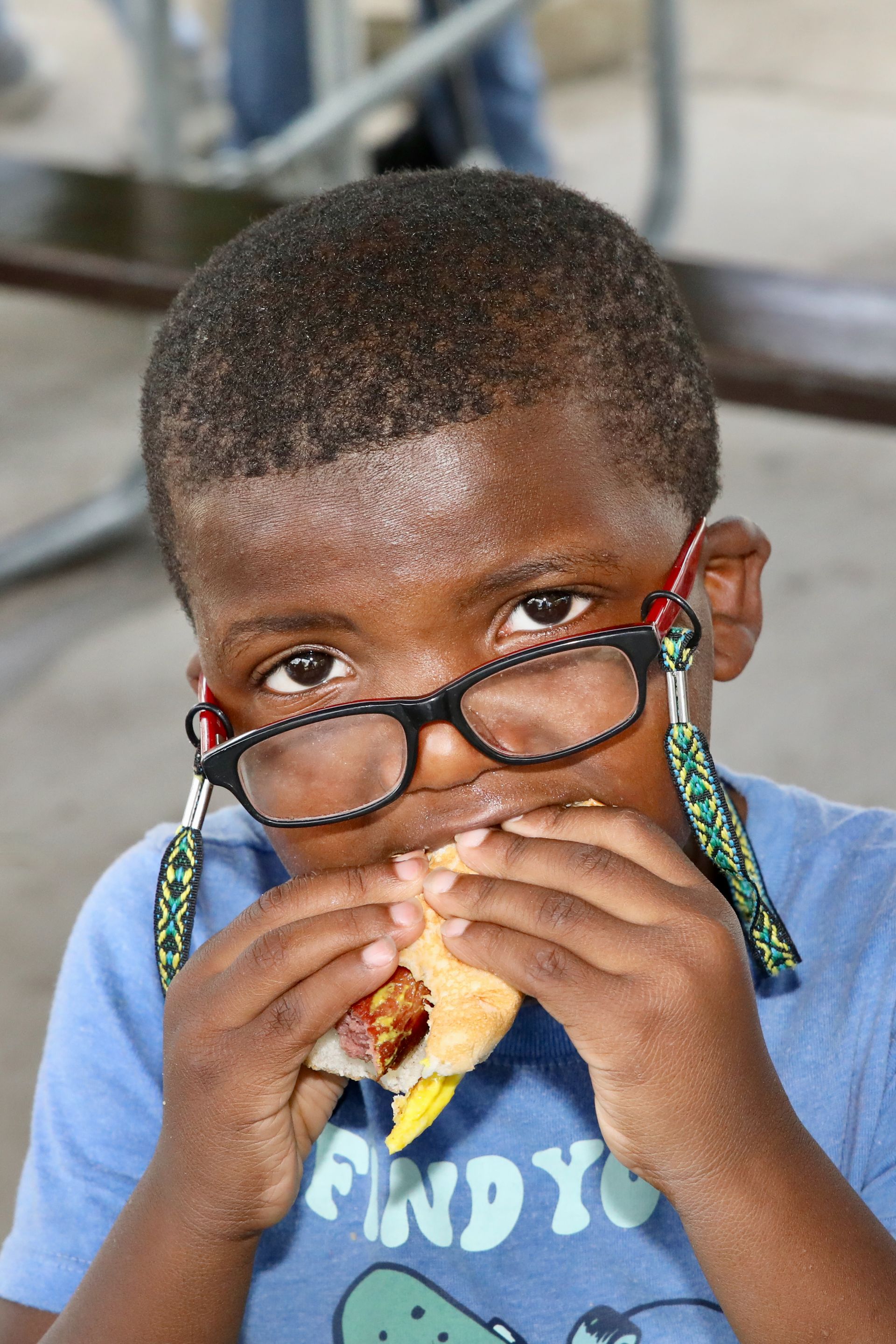 Boy eating a sandwich, wearing glasses with a strap. Outdoors, focused expression, holding food.