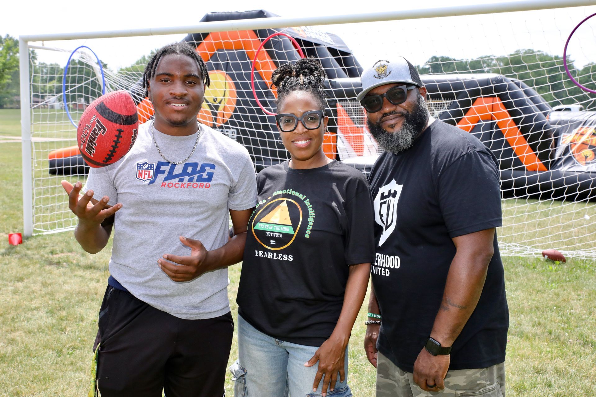 Three people standing in front of an inflatable obstacle course; one holding a football.