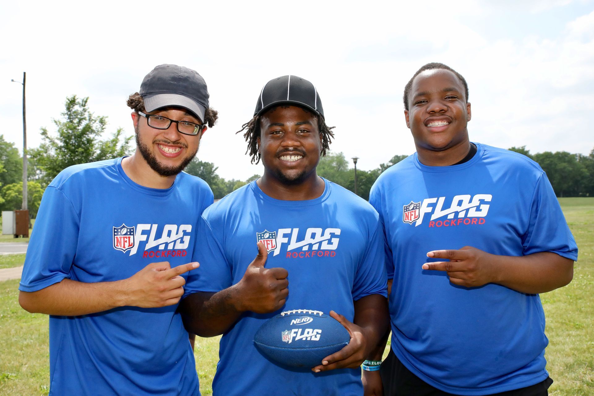Three people in blue NFL Flag Football shirts pose on a grassy field, holding a football.