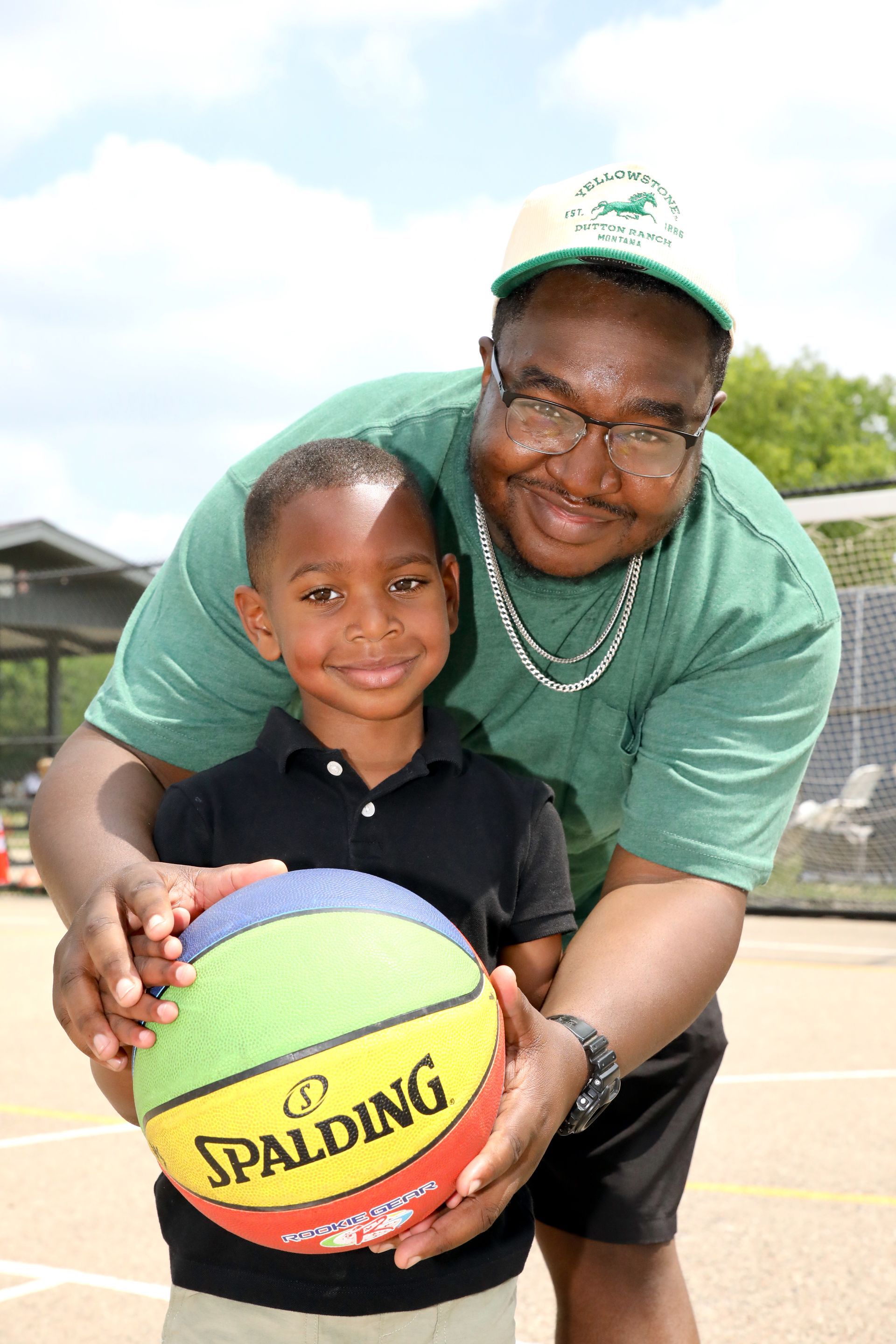 Man and child holding a colorful basketball outdoors. Man wears green shirt/hat. Child smiles.