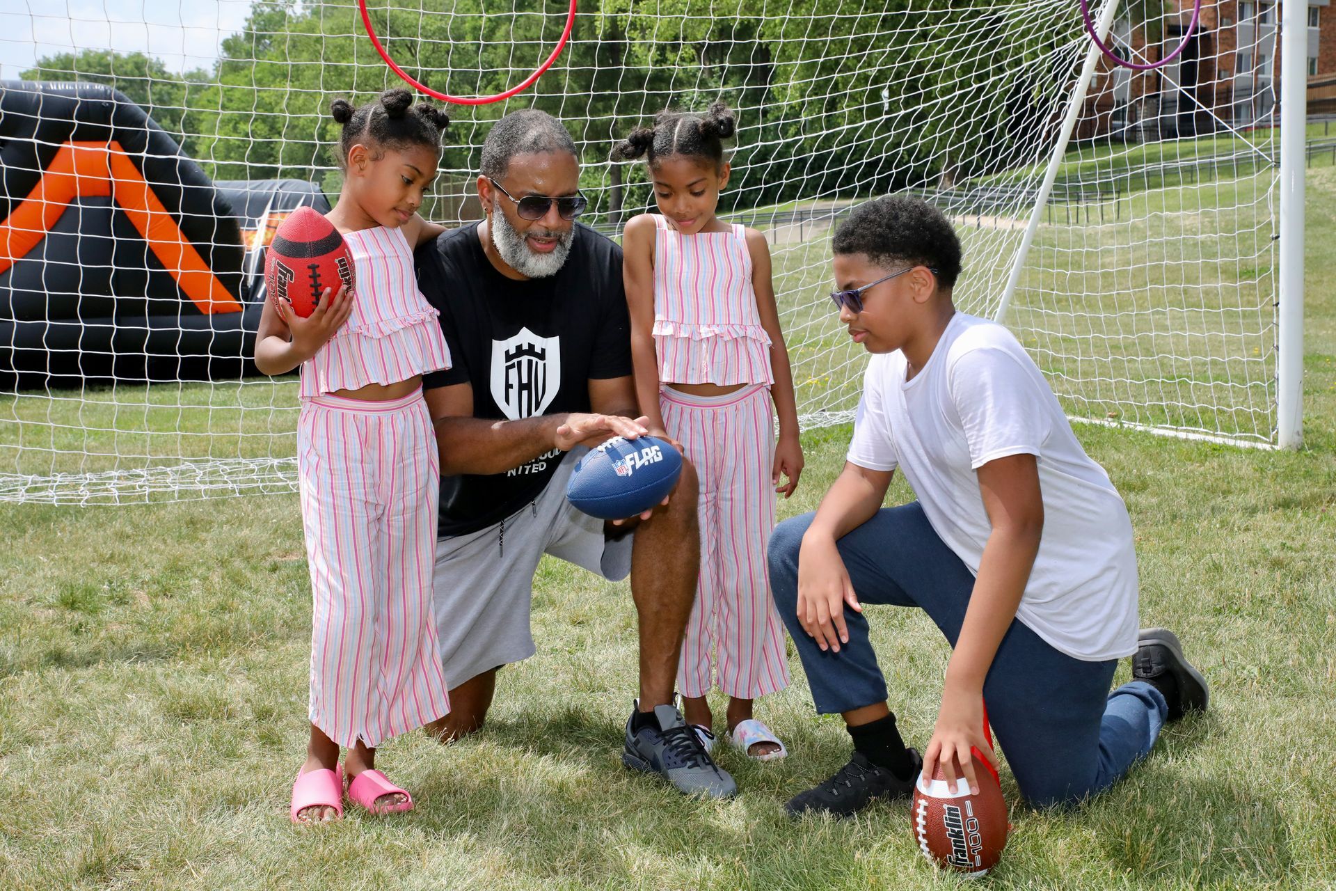 Family outdoors with footballs near a goal; two young girls in pink outfits, a man, and a boy in sunglasses.