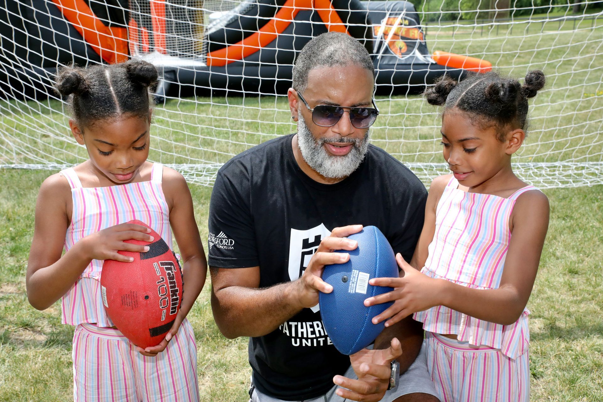 Man shows two young girls footballs outdoors. They are near an inflatable structure.