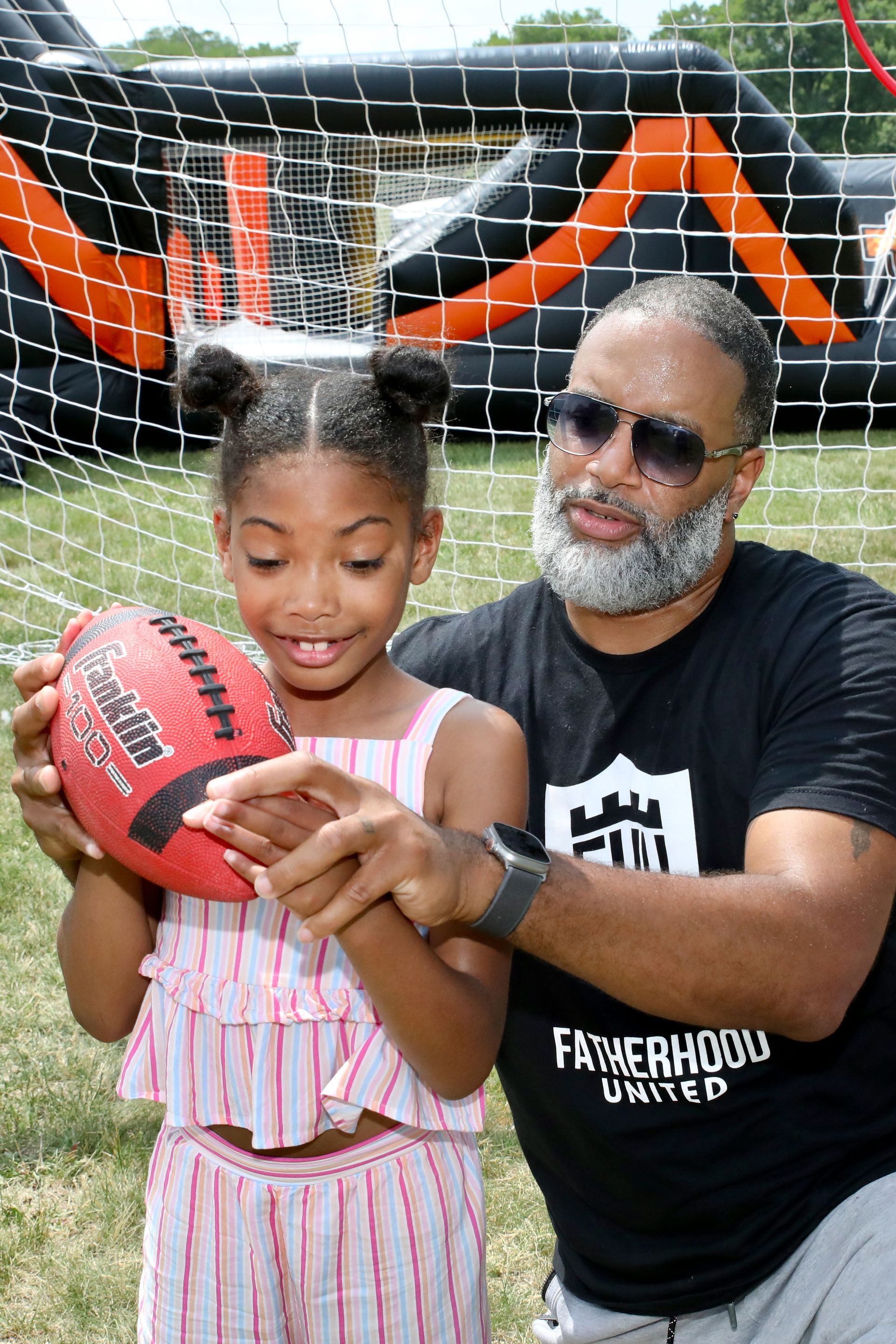 Man shows girl how to hold a football. Outdoors, bouncy castle background, sunny day.