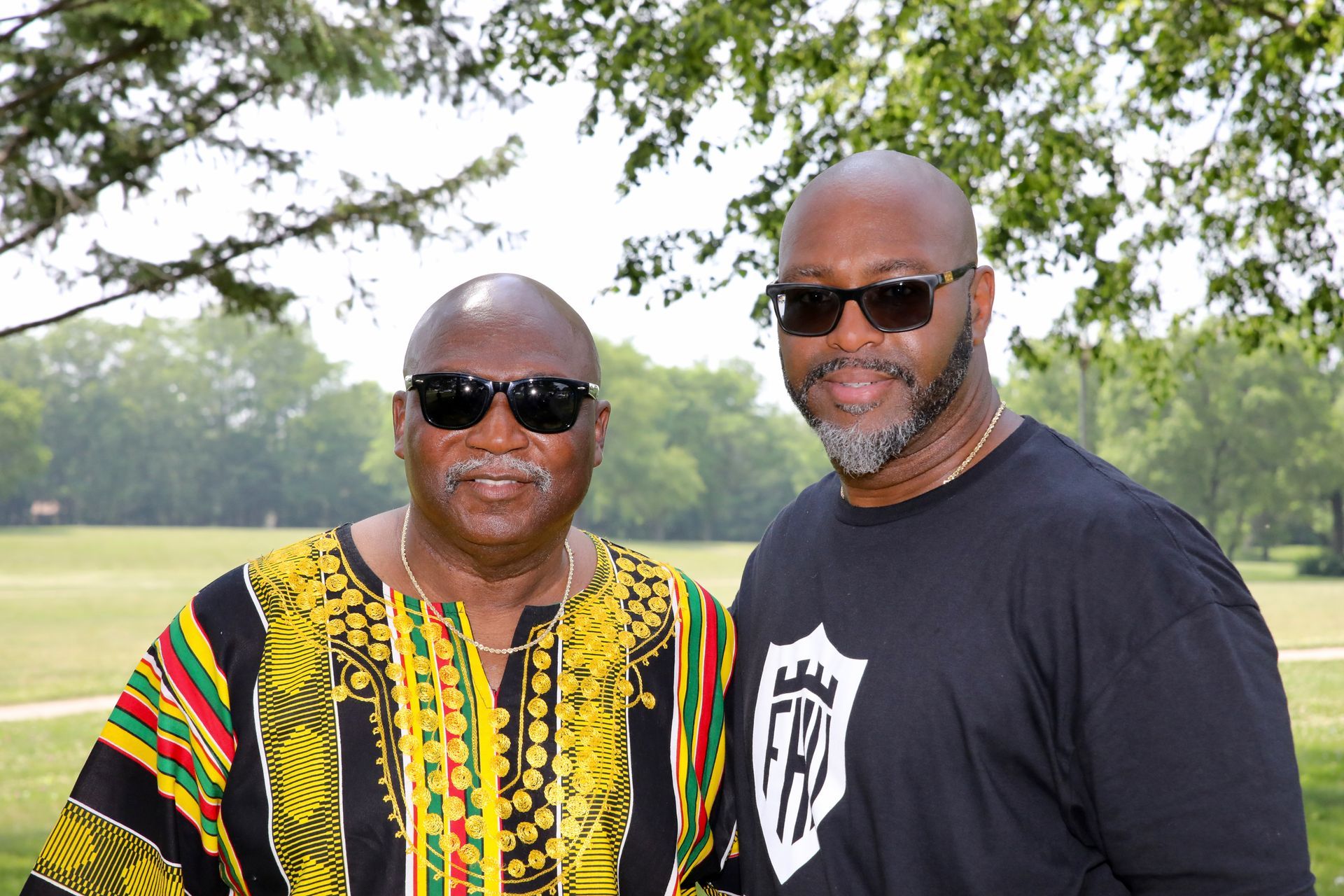 Two men wearing sunglasses, smiling. One in a colorful patterned shirt, other in a black shirt with a shield emblem. Outdoors, park setting.