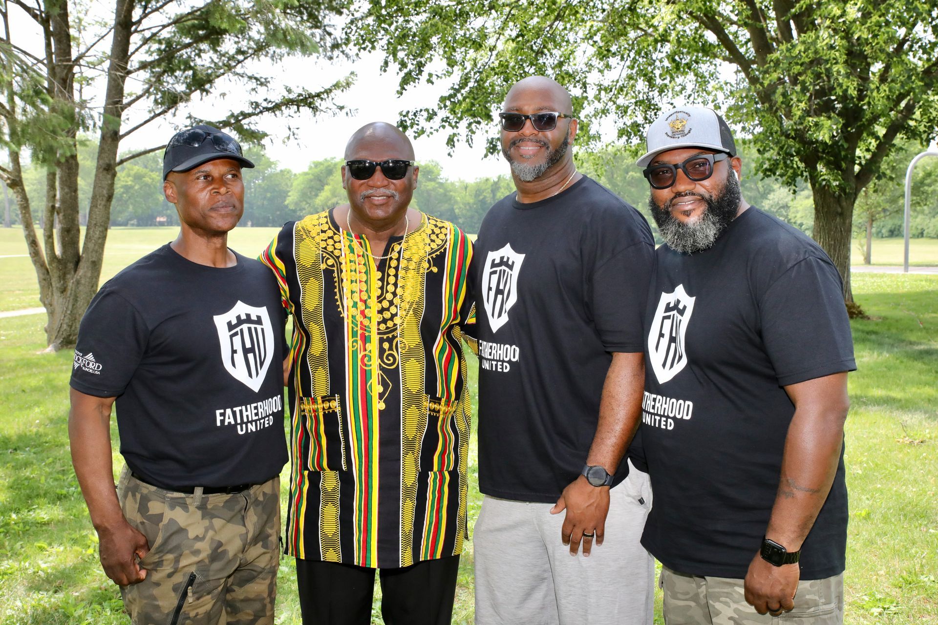 Four men pose outdoors. One wears patterned shirt, three wear matching black shirts with logo.