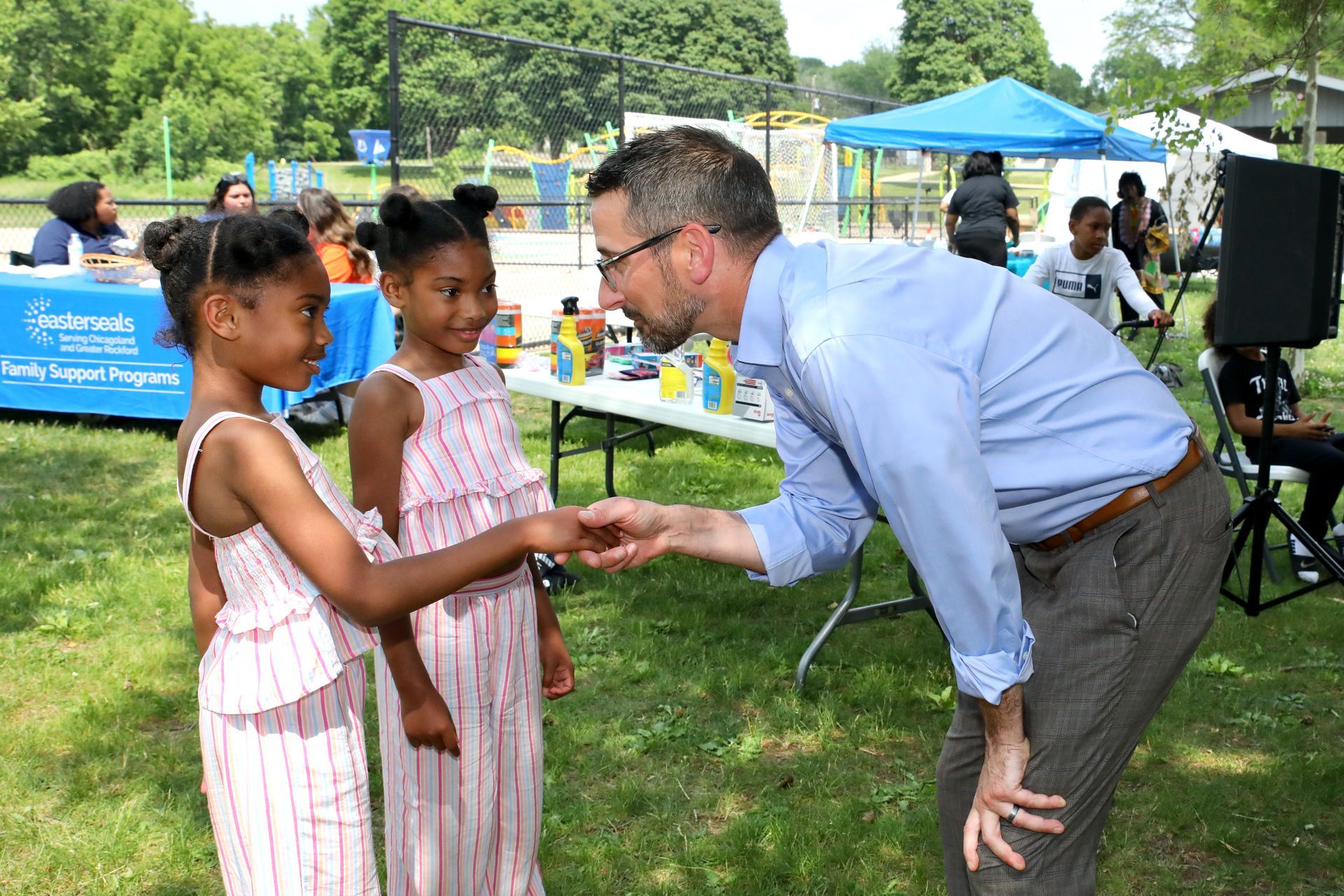 Man shaking hands with two girls outdoors at an event, with tables and other people in the background.