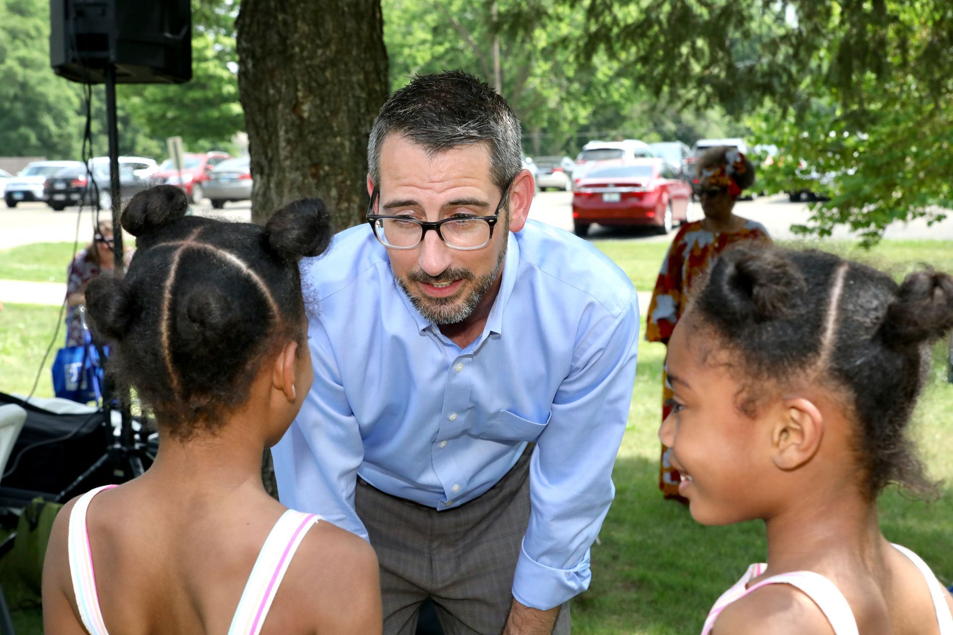 Man in glasses talking to two young girls with their hair in buns outside.
