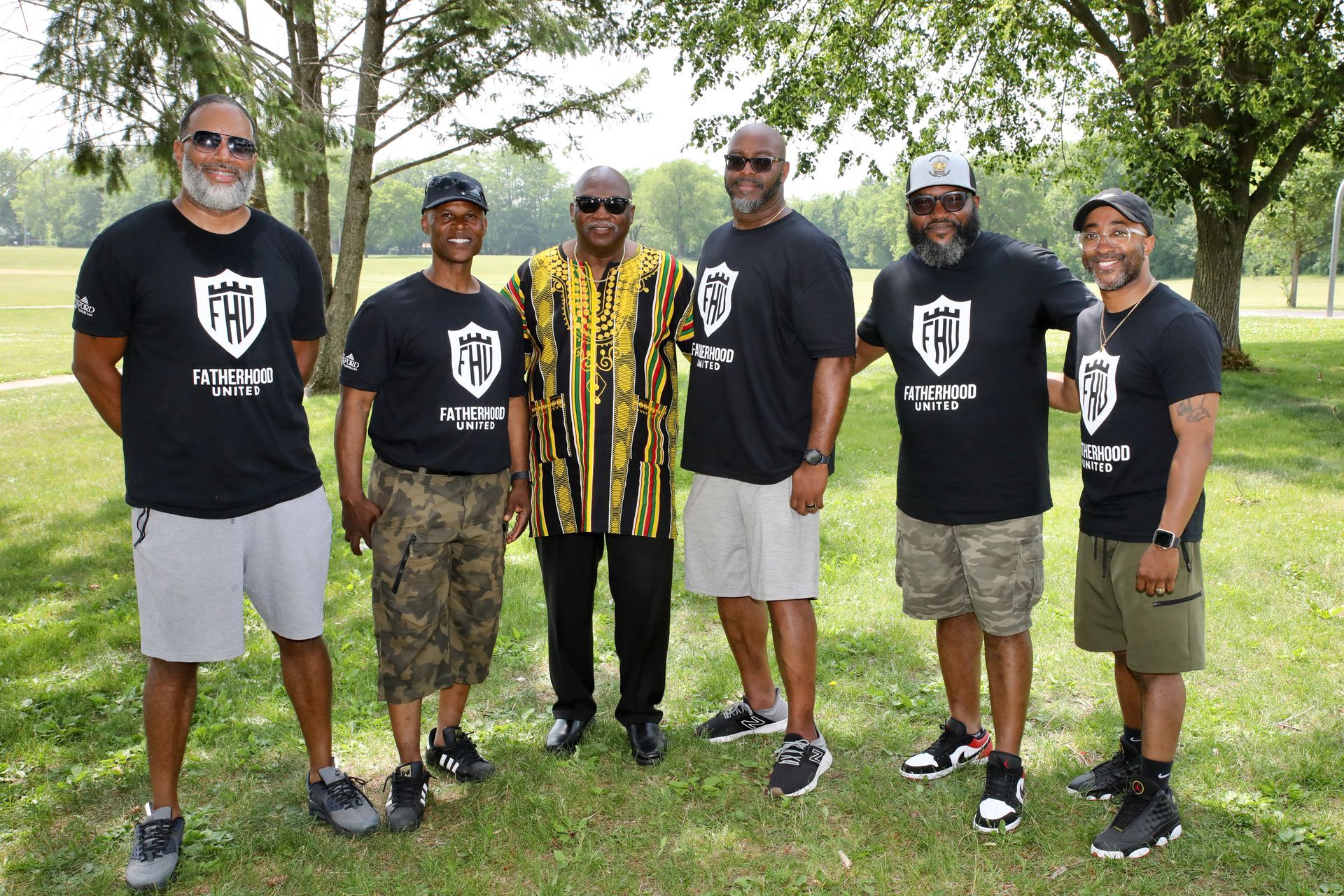 Six men standing outside, some wearing matching black shirts with a shield logo. One man wears a patterned shirt. Green space in background.