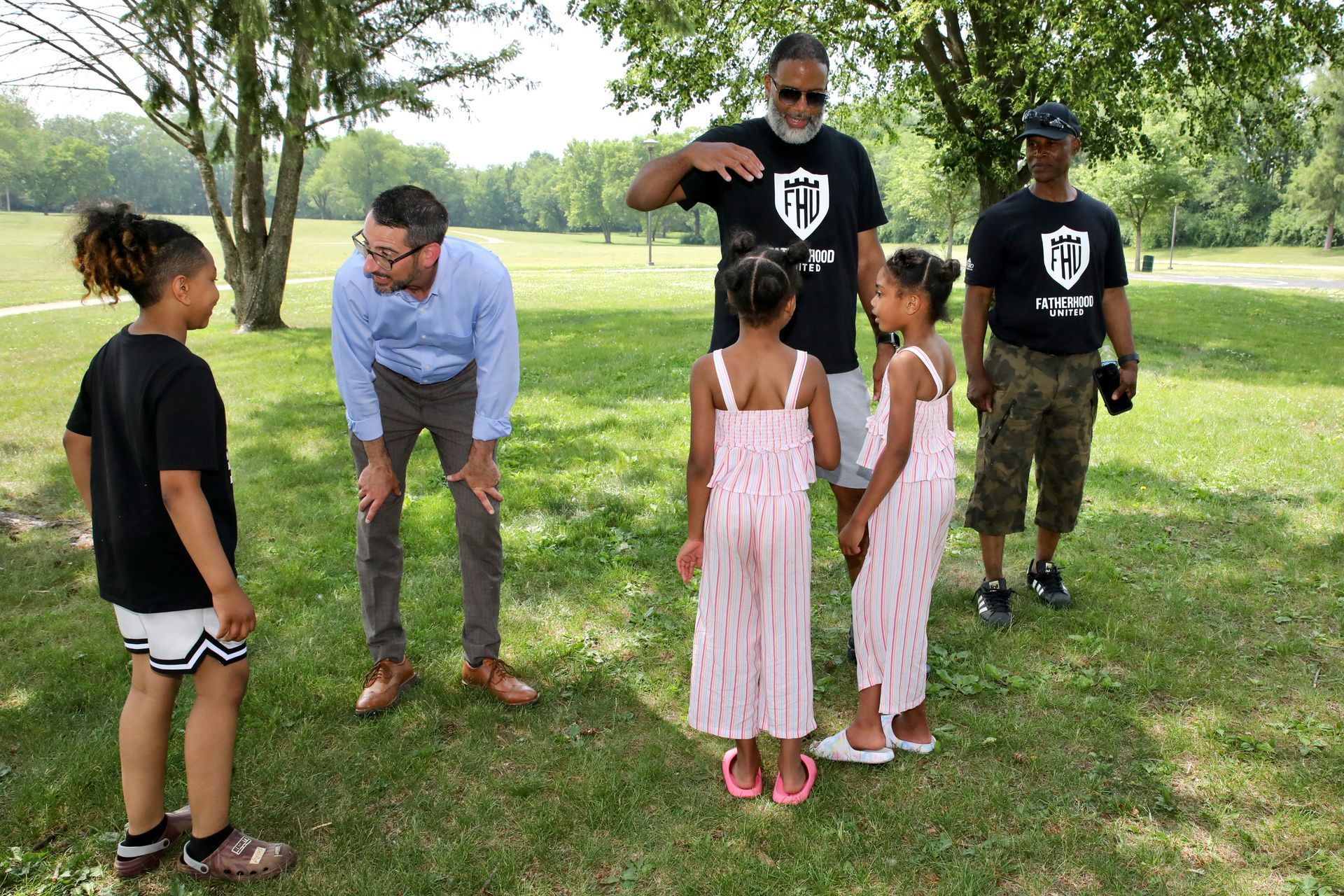 People in a grassy park, interacting. Man in blue shirt kneels, children in dresses stand. Others wear black shirts.