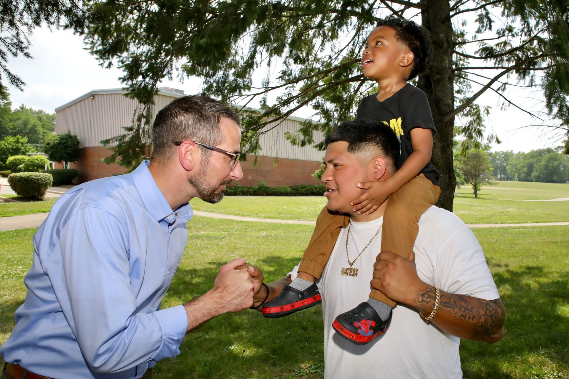 Man shakes hands with another man, who has a child on his shoulders, in a park.