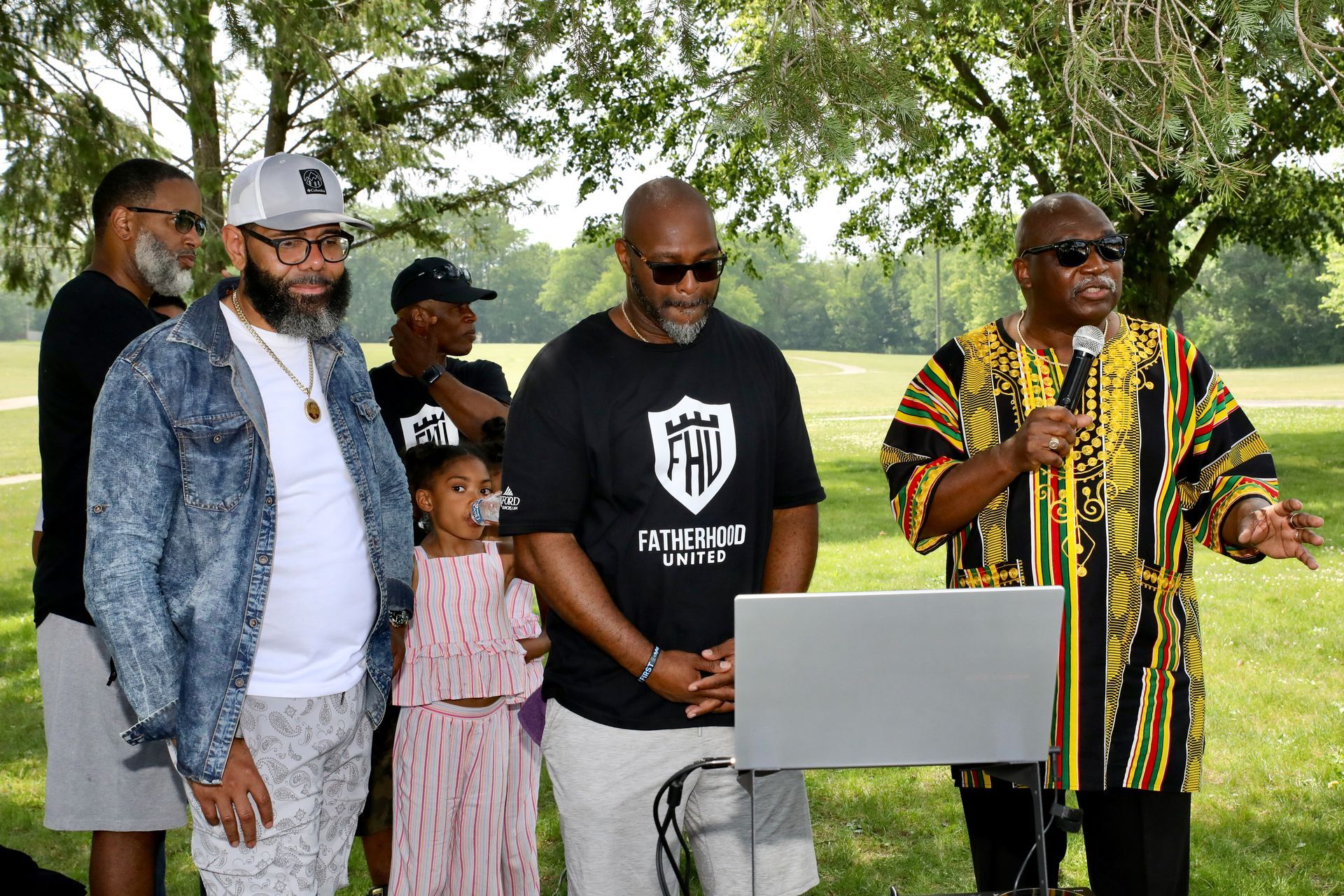 People gathered outdoors, one speaking into a mic; others stand nearby near a laptop.
