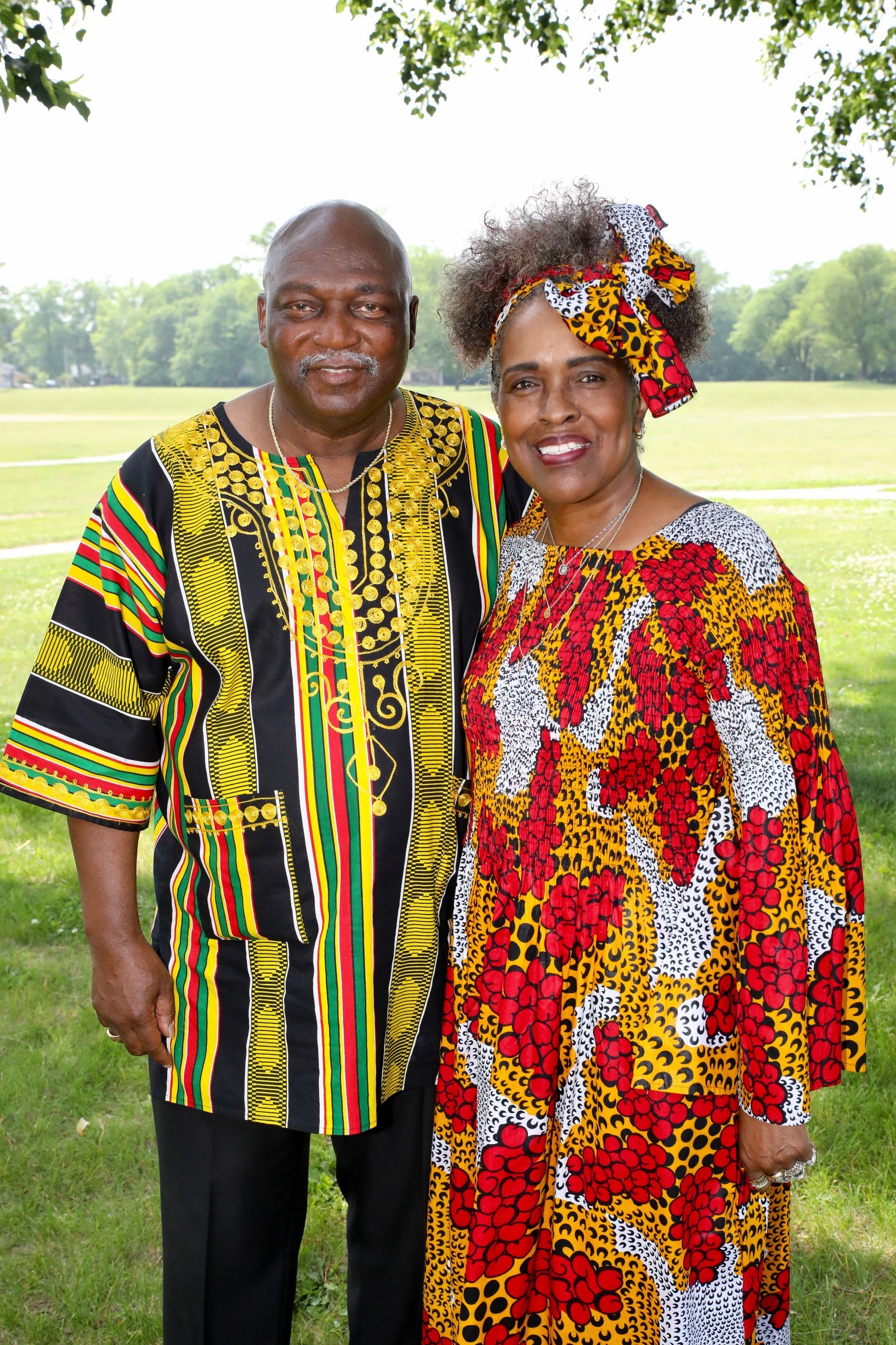 Couple in African-print attire poses outdoors; man in yellow shirt, woman in red dress, both smiling.