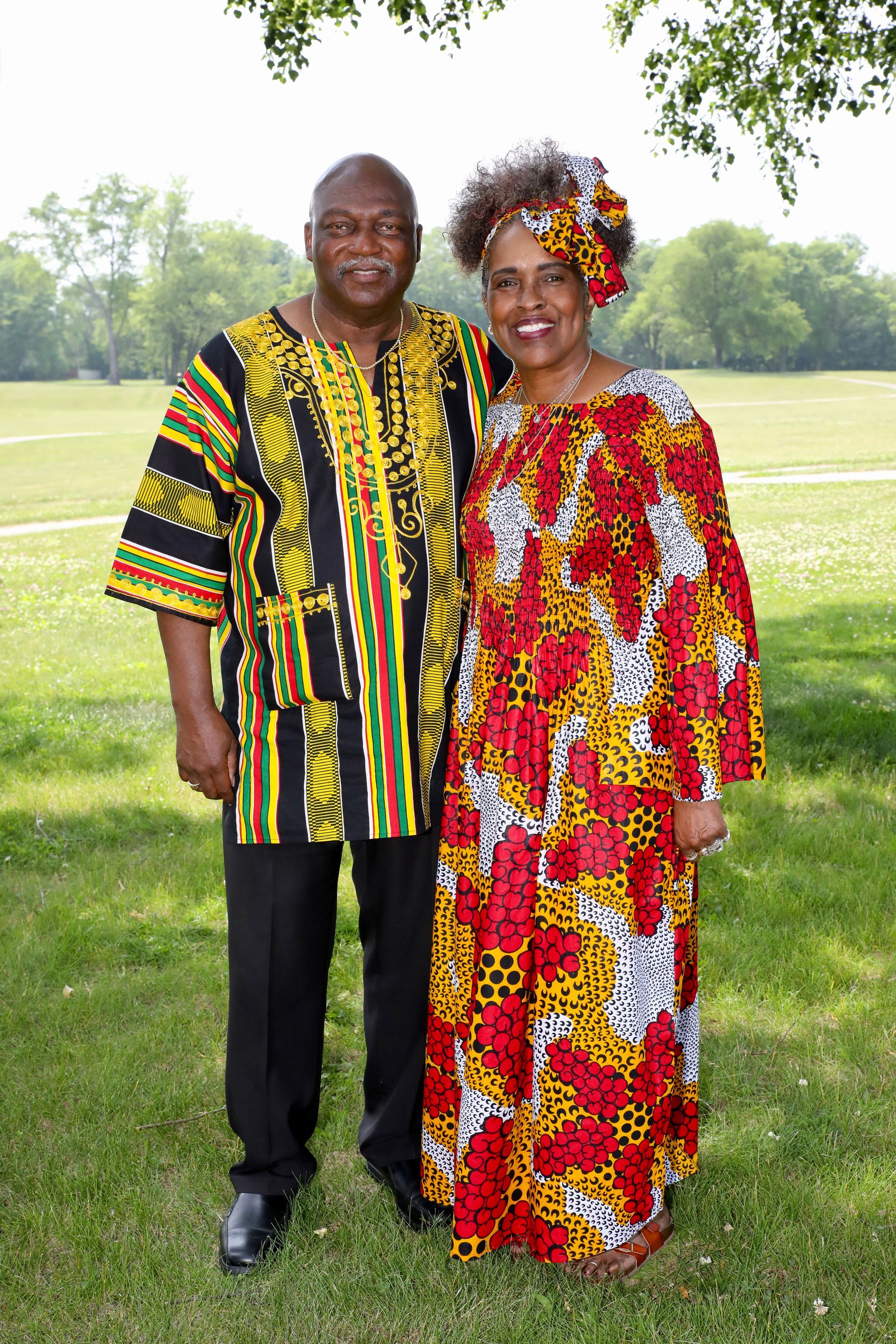 Couple standing outdoors, wearing colorful African attire, smiling.