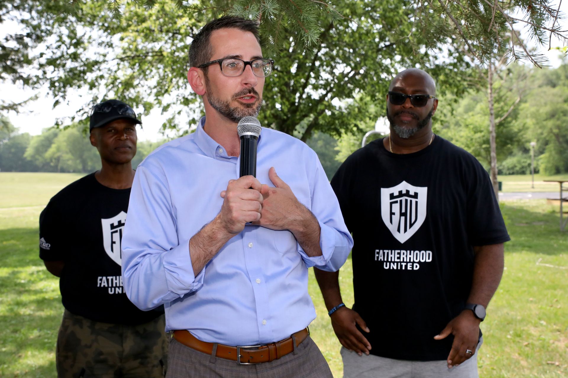 Man speaking into microphone, two men stand behind him; all wear 