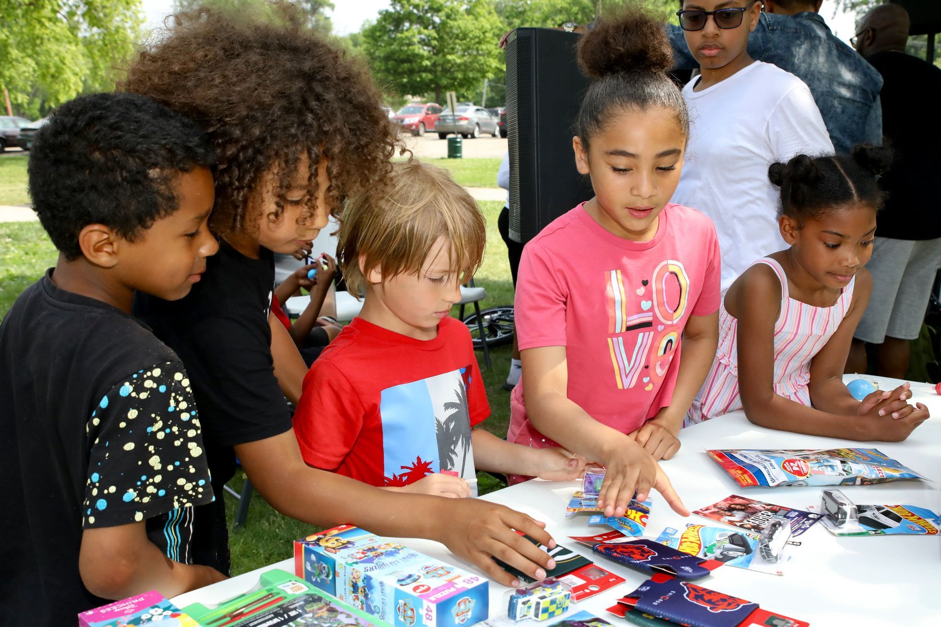 Children looking at items on a table outside. Various skin tones, excited expressions. Bright colors and sunny setting.
