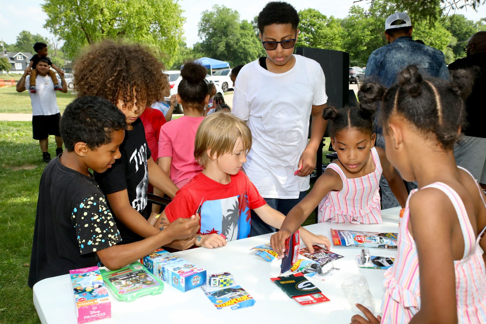 Children at an outdoor table selecting toys on a sunny day.