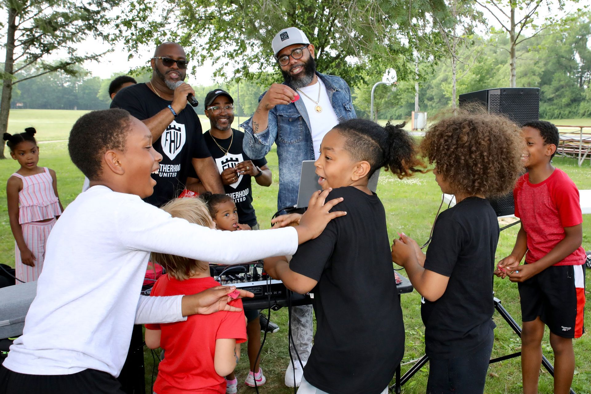 Children dancing outdoors with adults near a sound system. Green grass, trees, and cloudy sky in background.