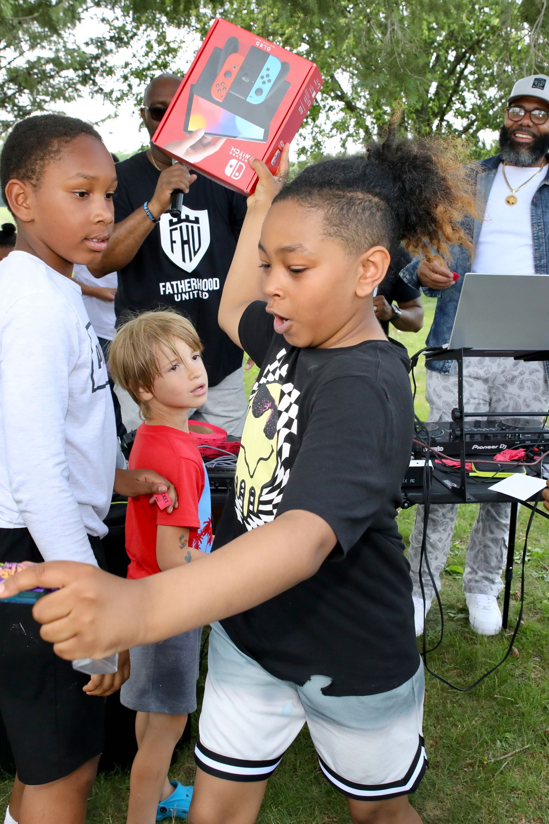 Boy holding up a red gift box, possibly a game console, outdoors. Others watch nearby.