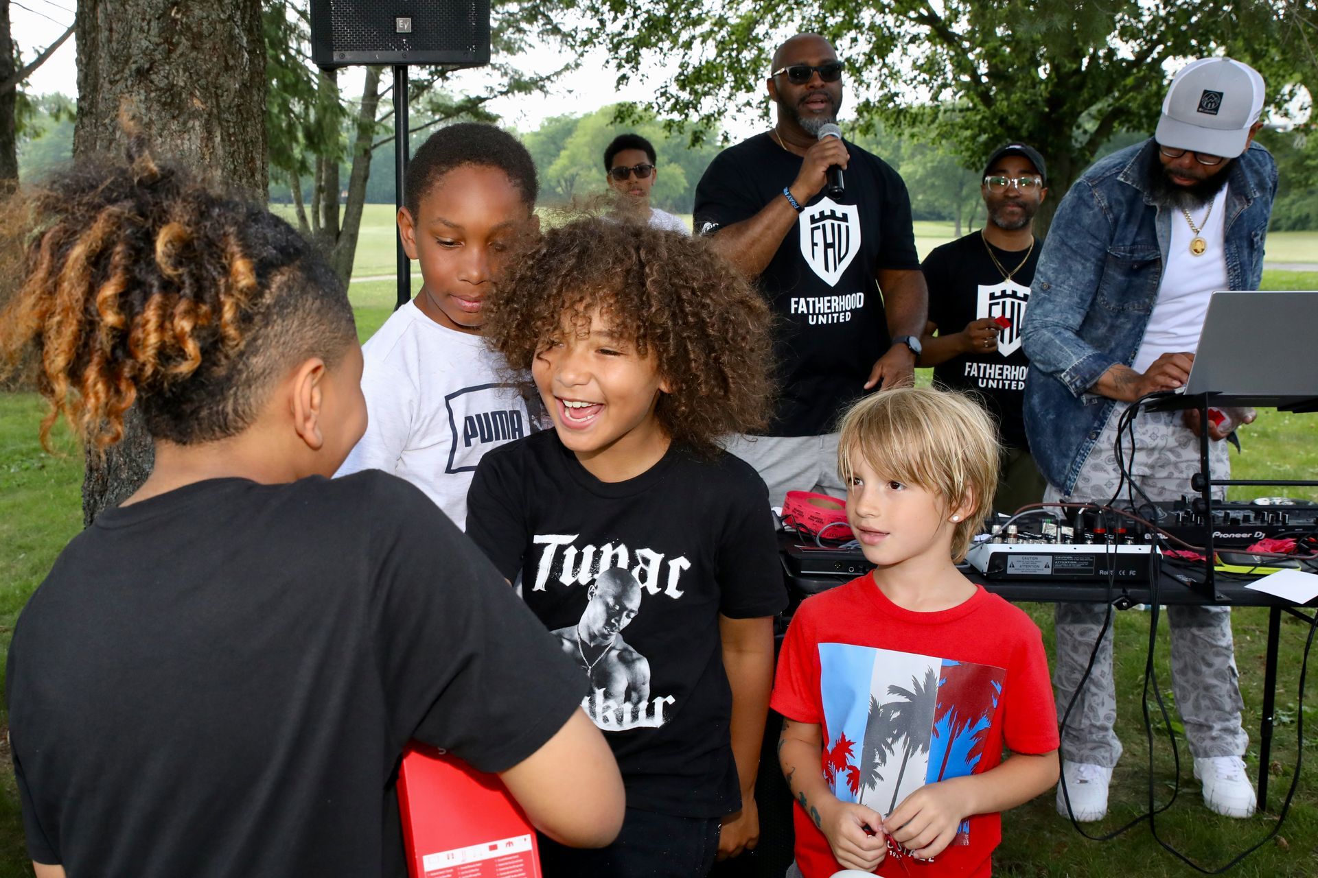 Group of people outdoors with children at an event, speaker and DJ set up.