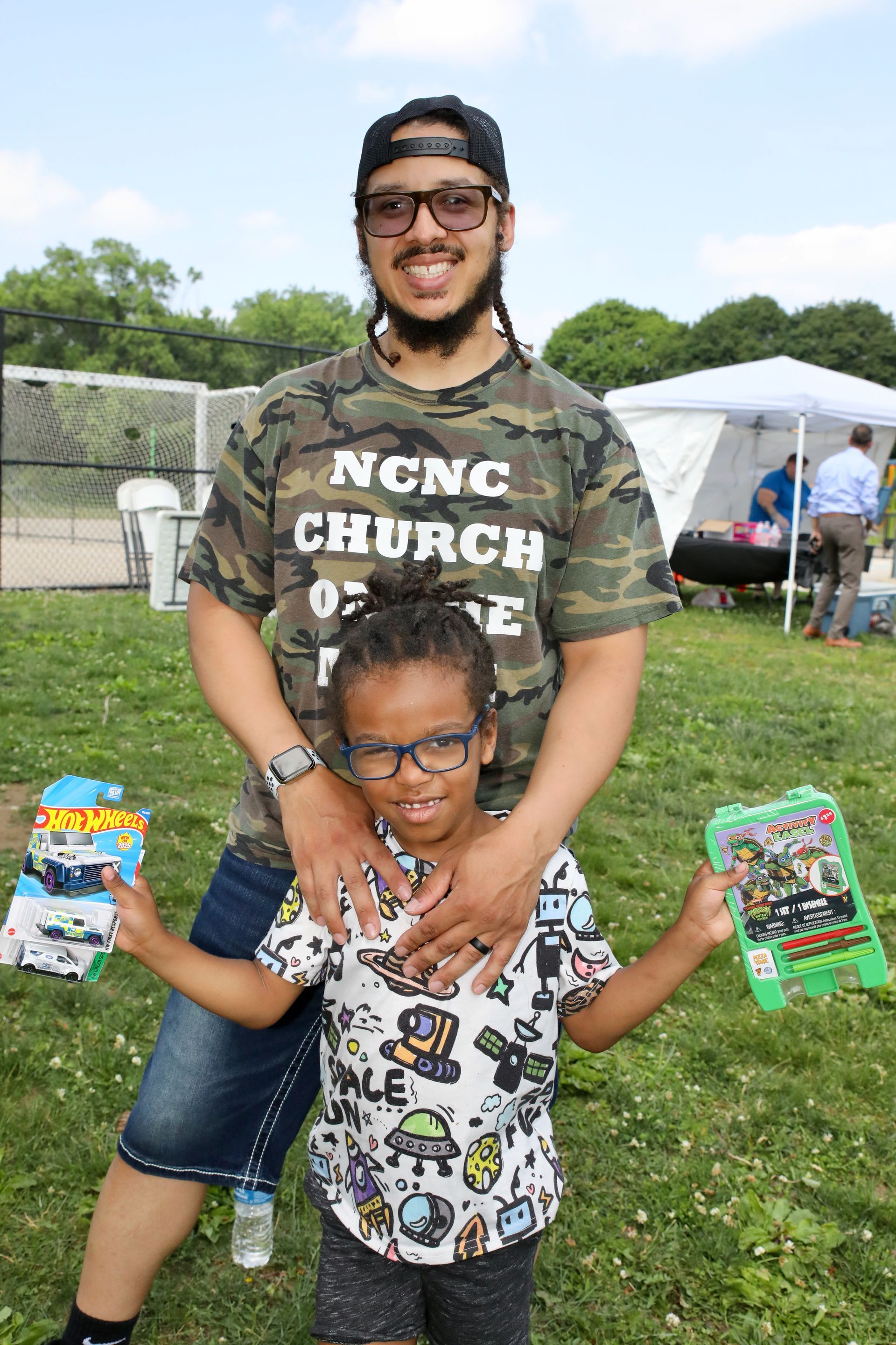 Man and child smiling, holding toys outdoors. Man in camo shirt, child in patterned shirt, both wearing glasses.
