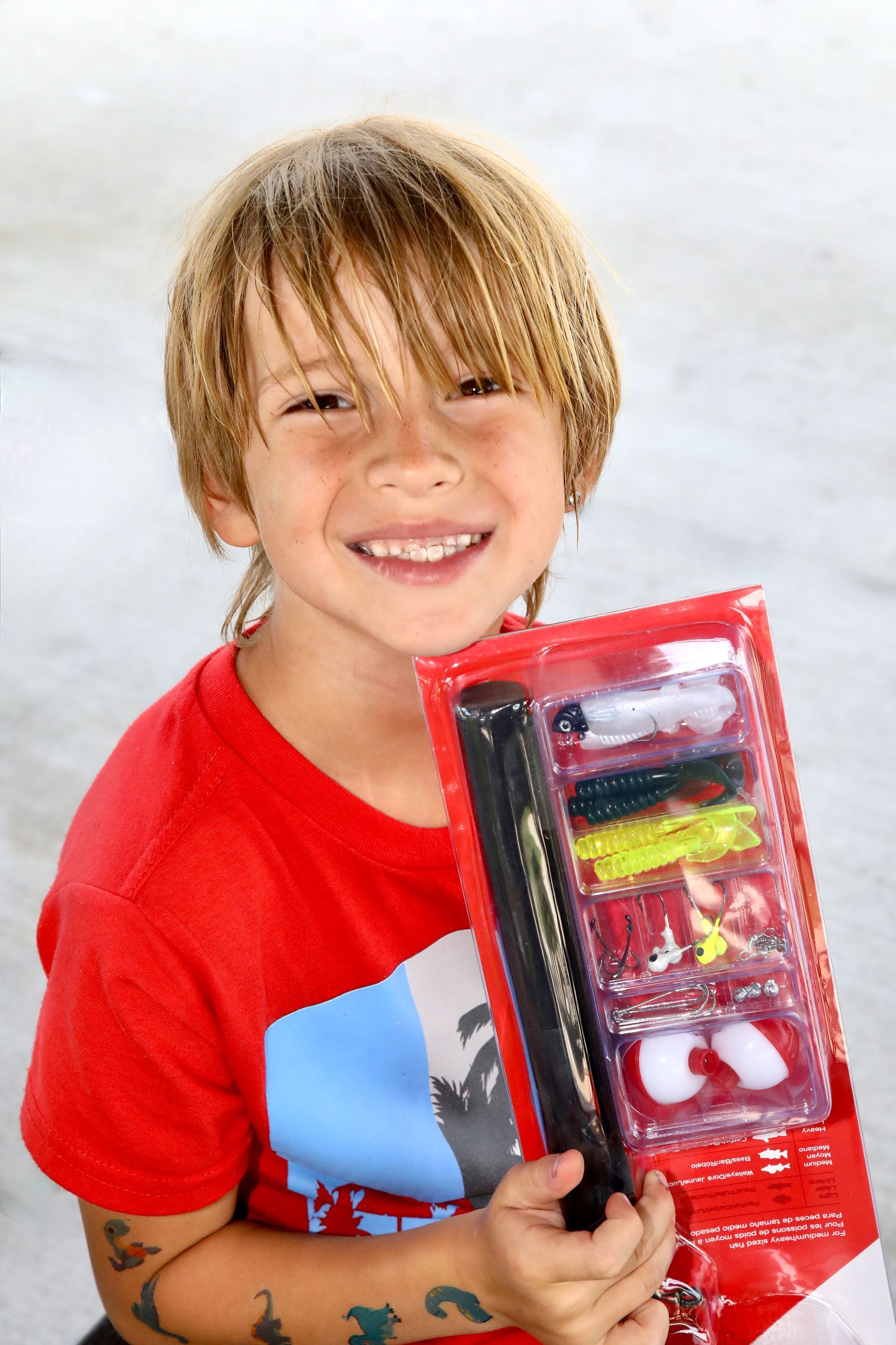 Boy with shoulder-length hair smiles, holding a fishing lure kit. He wears a red shirt.