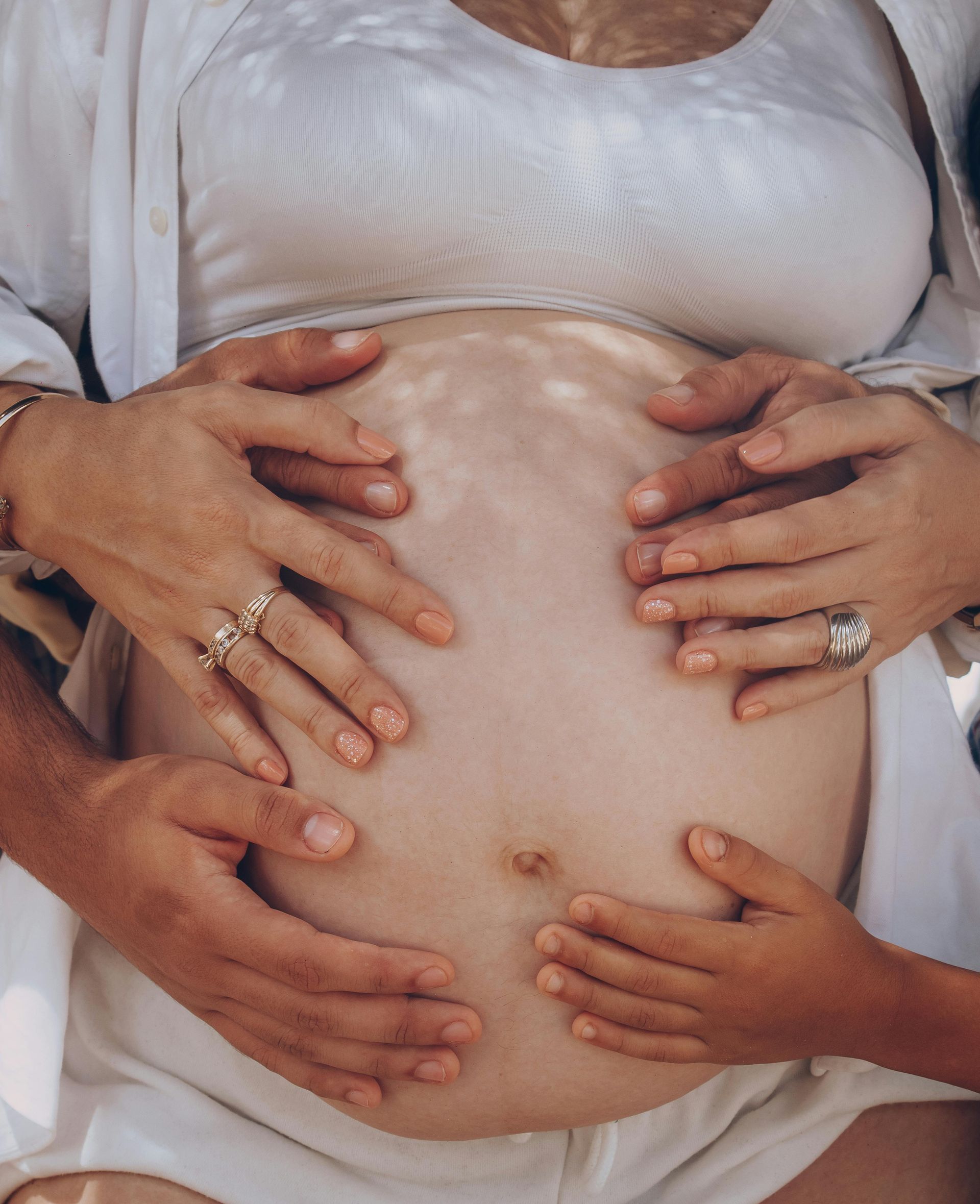 Pregnant belly with four hands caressing it, wearing rings, in a white outfit.