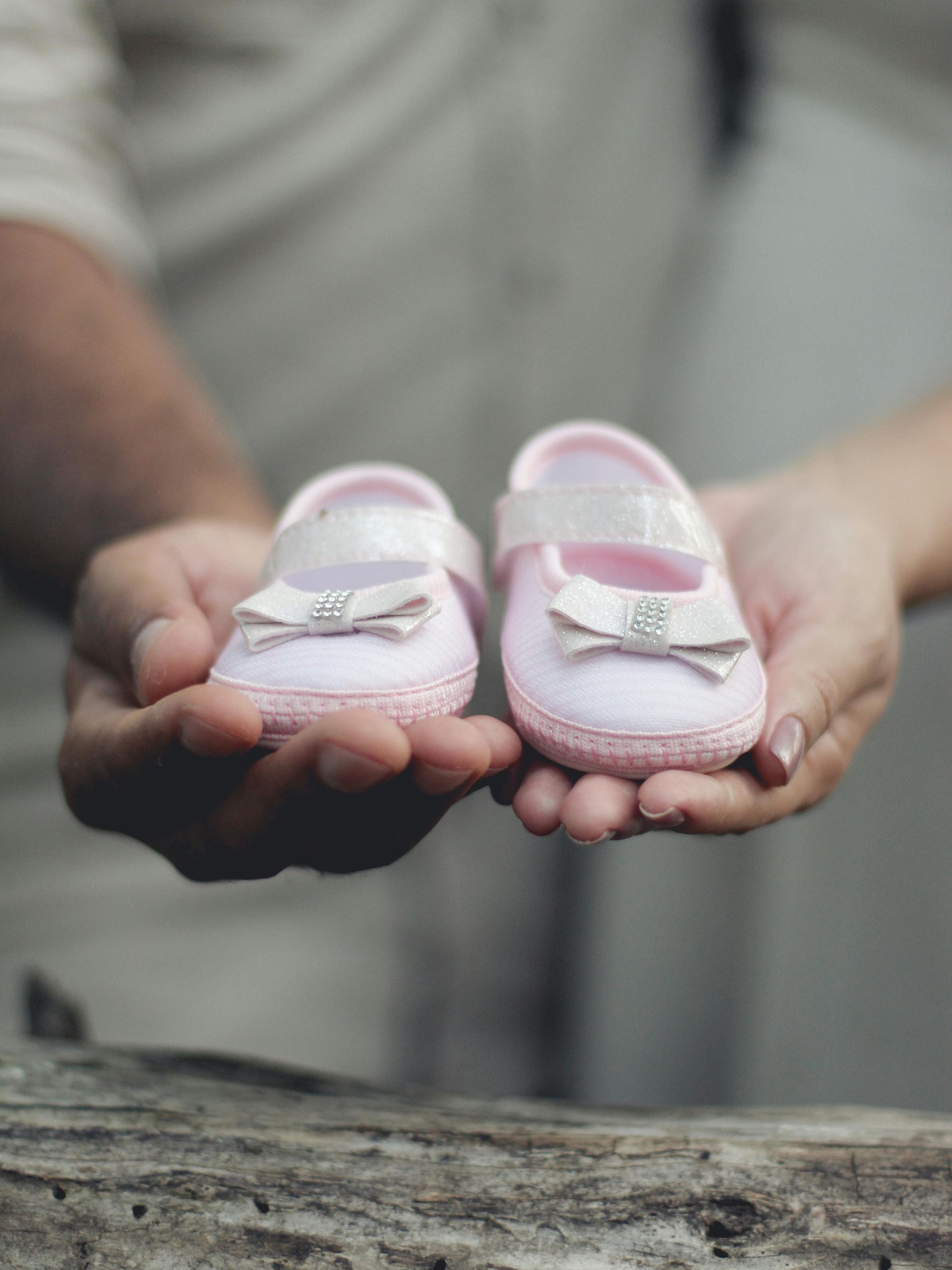 Hands holding two pink baby shoes with bows.