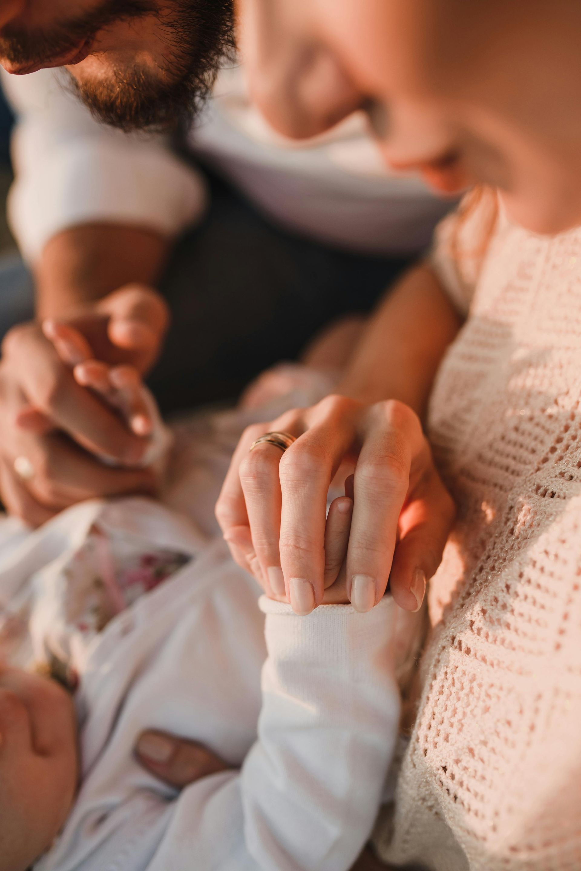 Parents holding the tiny hand of their baby, close-up. Soft lighting, focus on hands and baby in white.