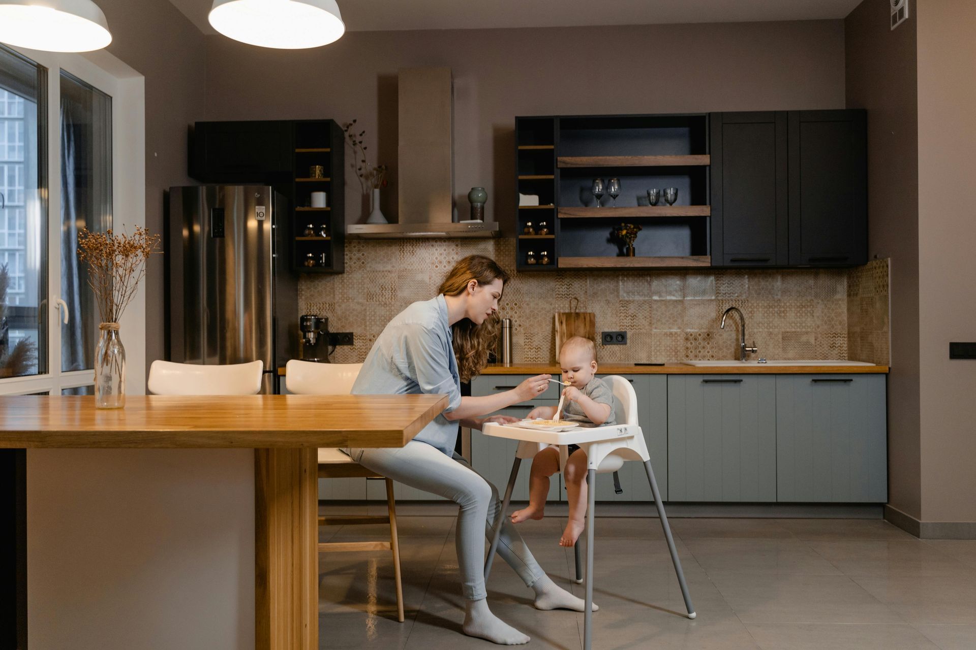 Woman feeding a baby in a modern kitchen. She sits at a table near a high chair, both near cabinets and a window.