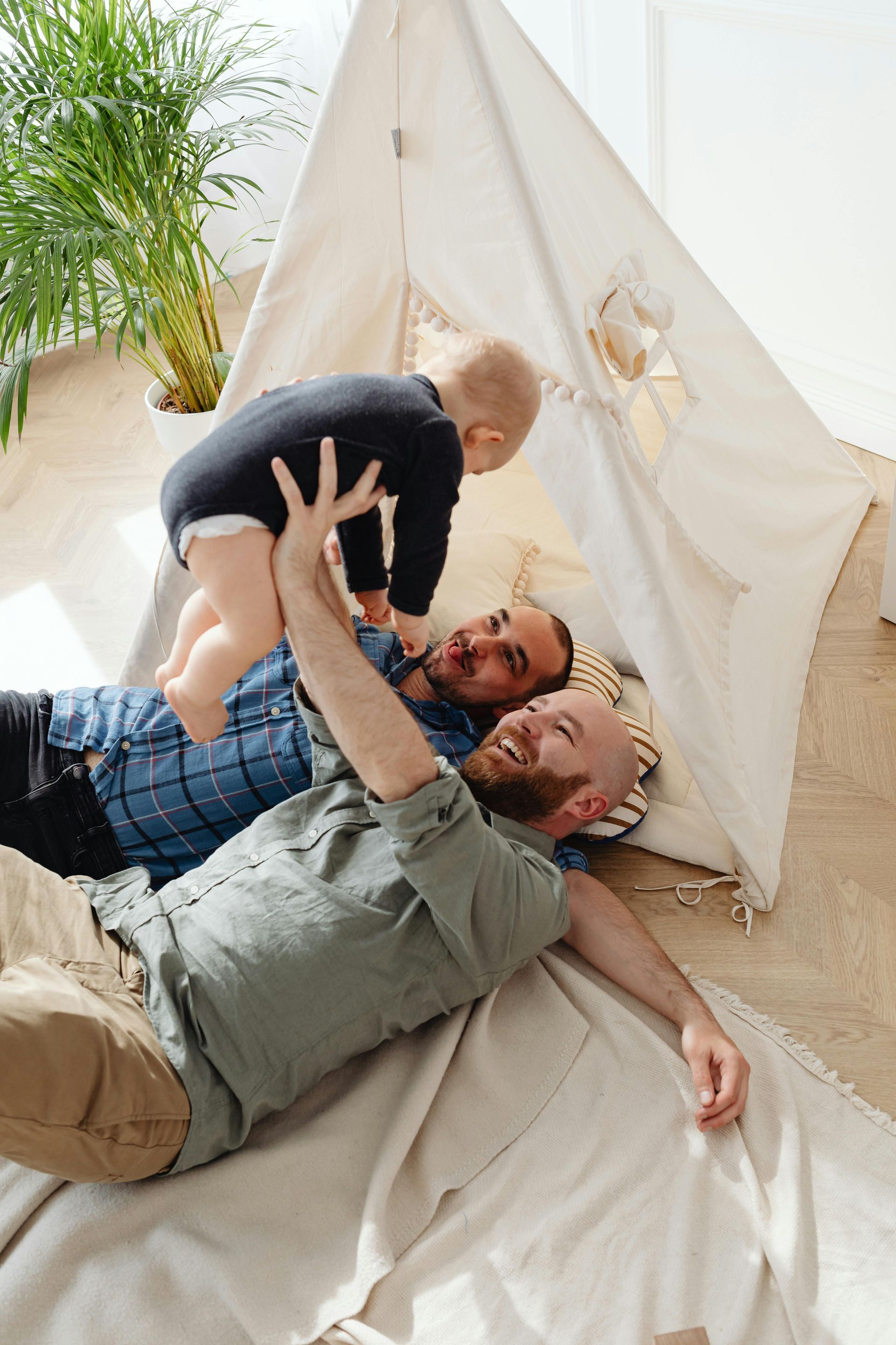 Two men and a baby playing in a teepee. One man lifts the baby in the air; all three are smiling.
