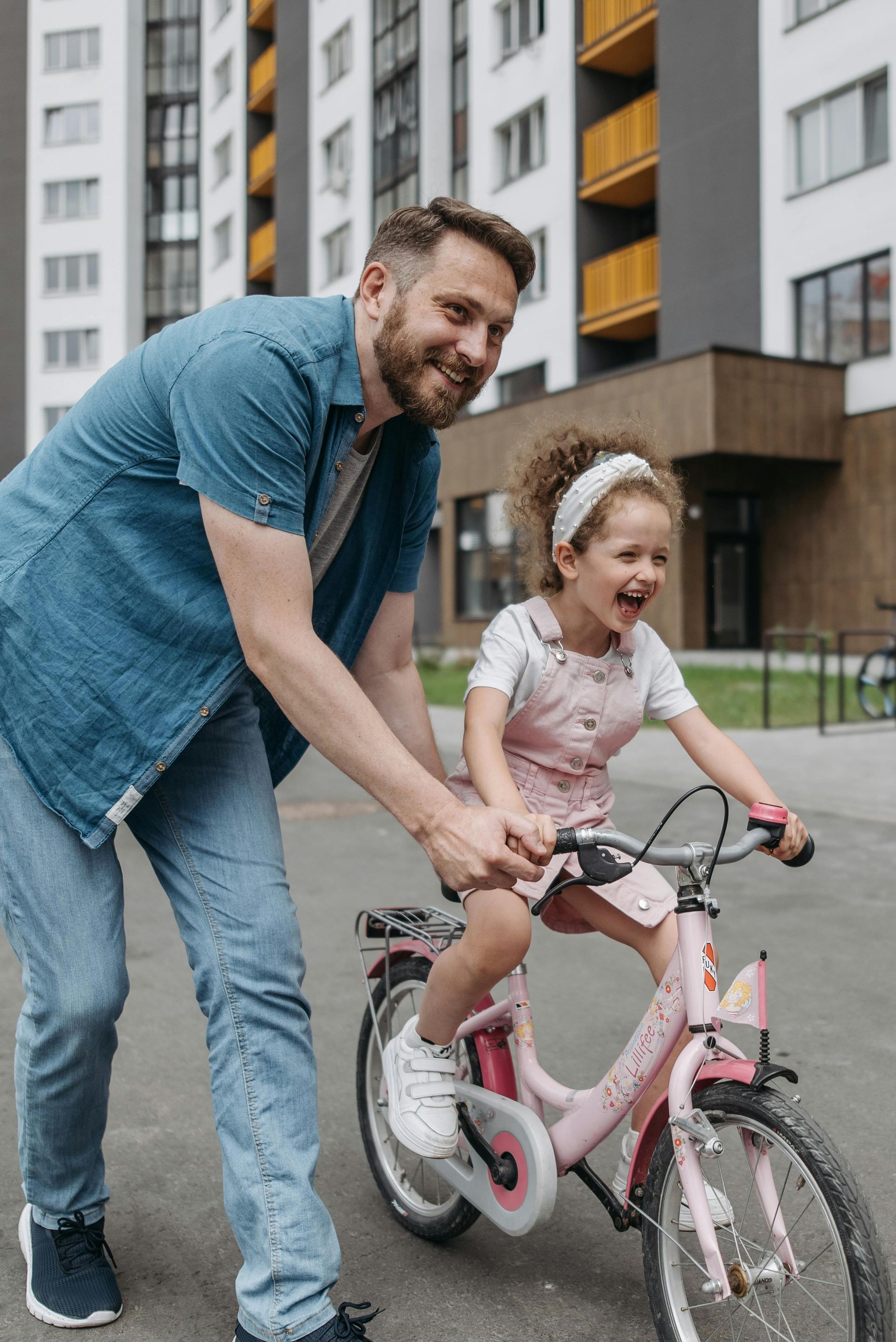 Father helps child learn to ride a pink bicycle, smiling, in front of an apartment building.