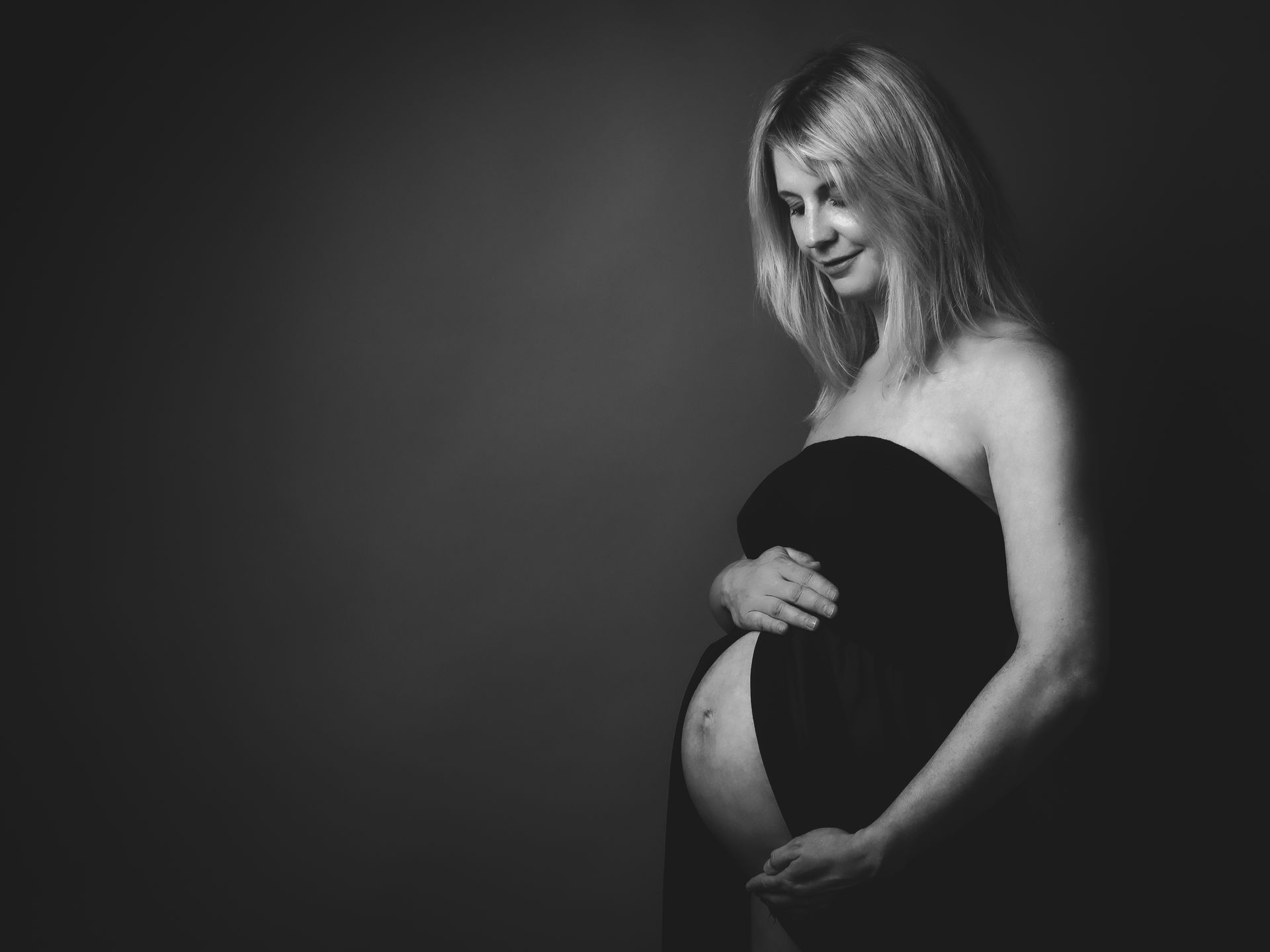 Schwangere Frau in einem schwarzen Kleid, lächelnd und den Bauch haltend, vor dunklem Hintergrund
