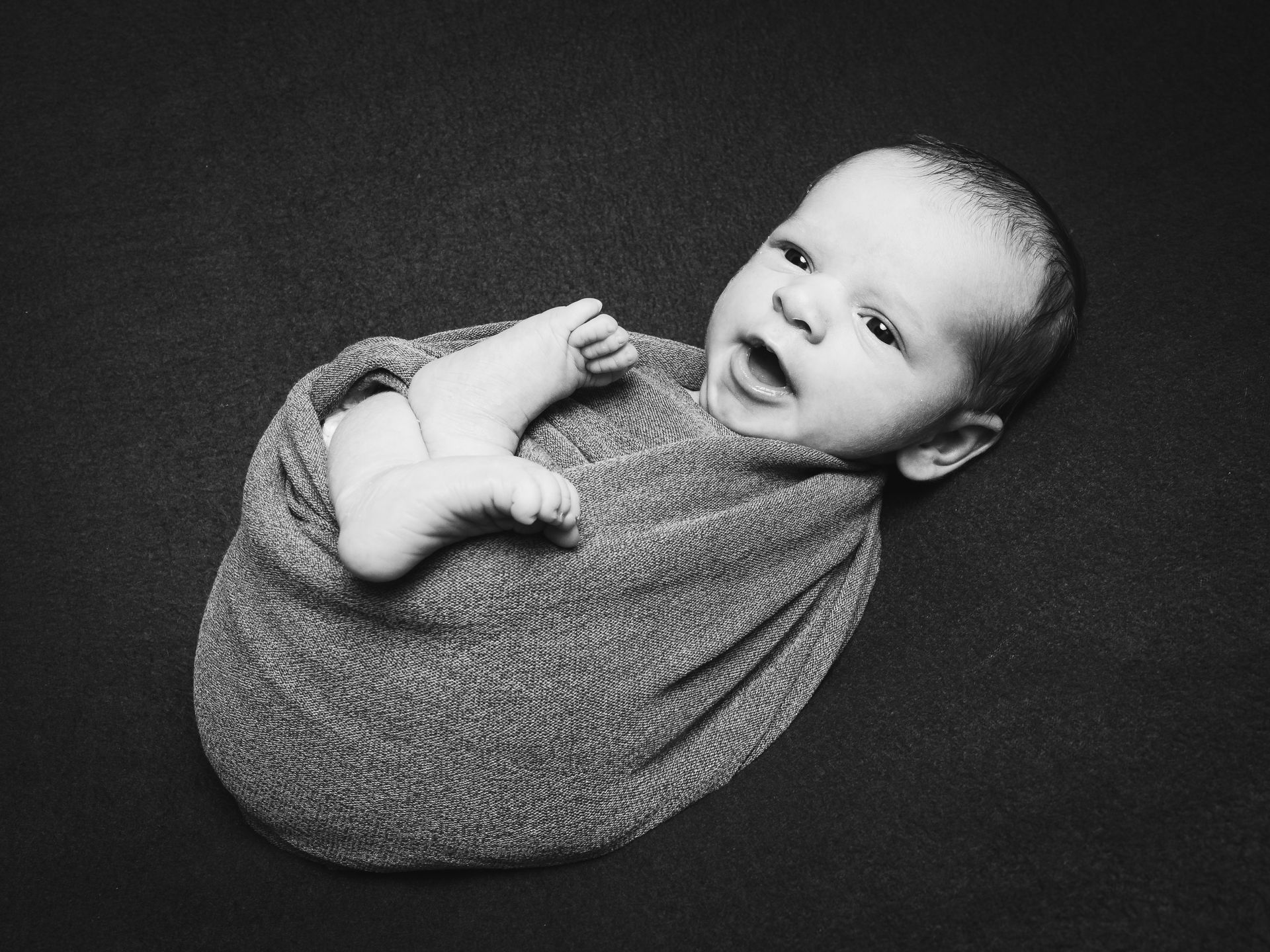Ein Baby, eingewickelt in eine strukturierte Decke, liegt vor dunklem Hintergrund und schaut in die Kamera.
