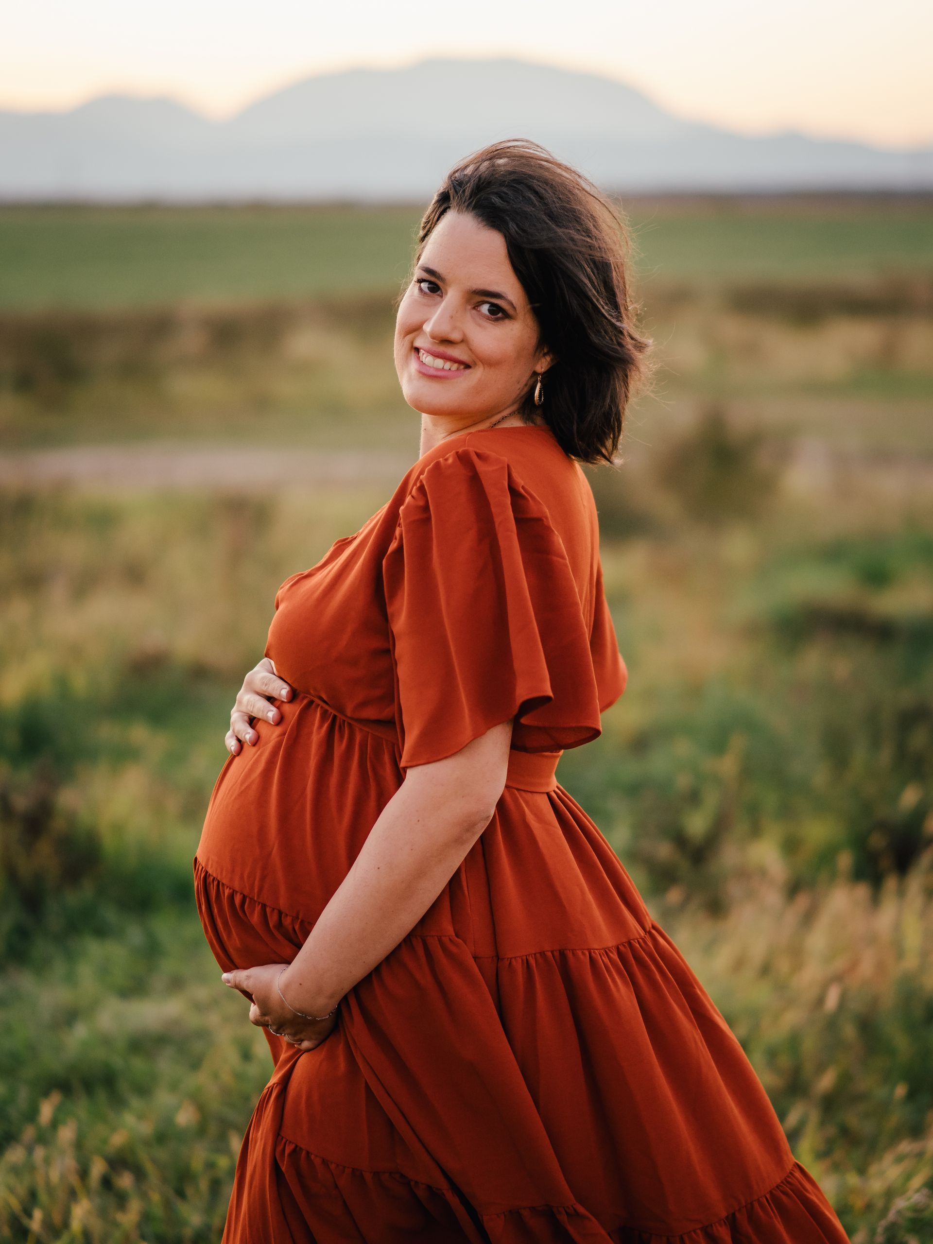 Schwangere Frau in einem rostfarbenen Kleid steht bei Sonnenuntergang im Freien und lächelt über die Schulter.