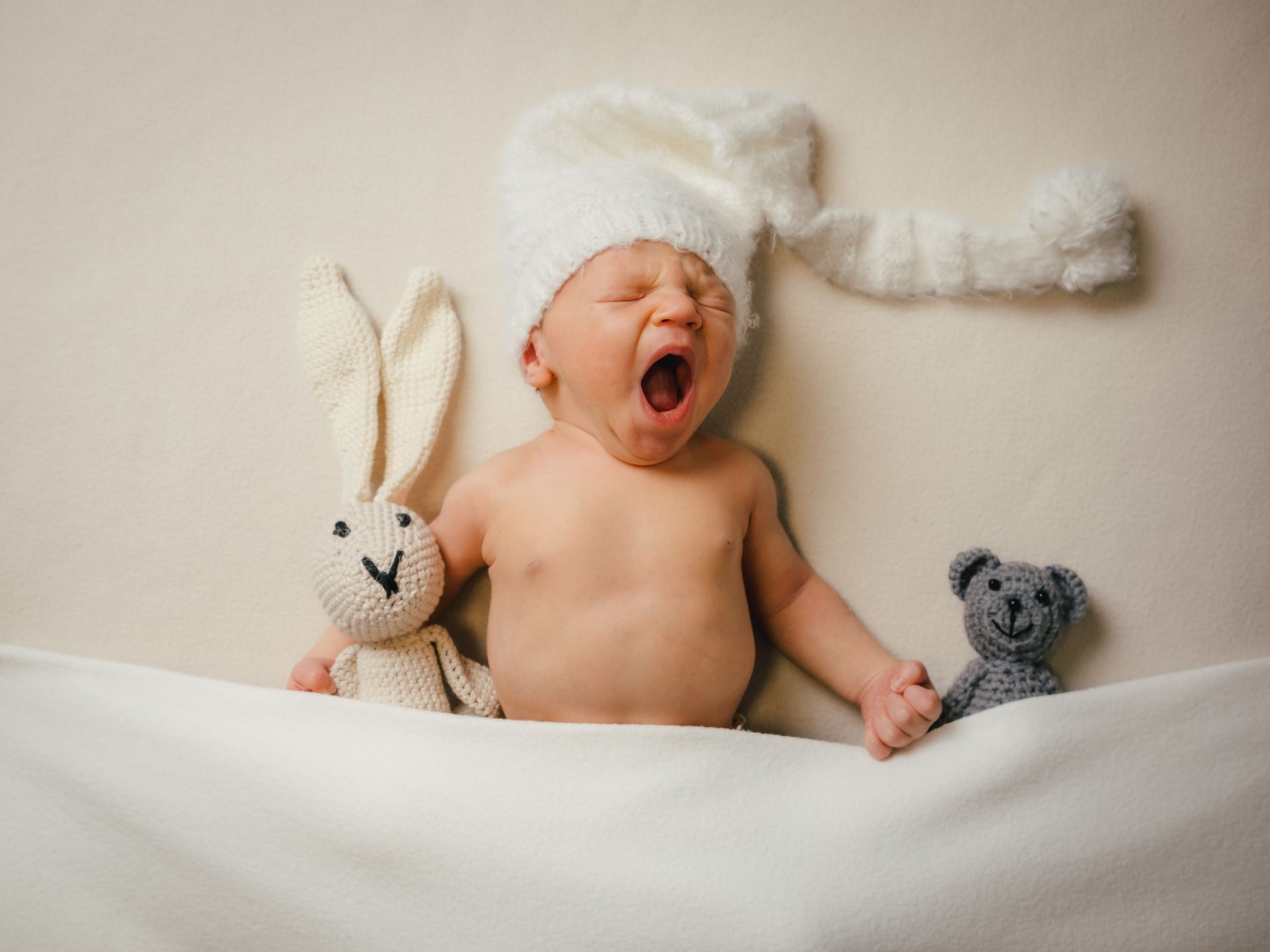 Ein gähnendes Baby mit weißem Hütchen, im Bettchen mit Plüschhasen und -bären.