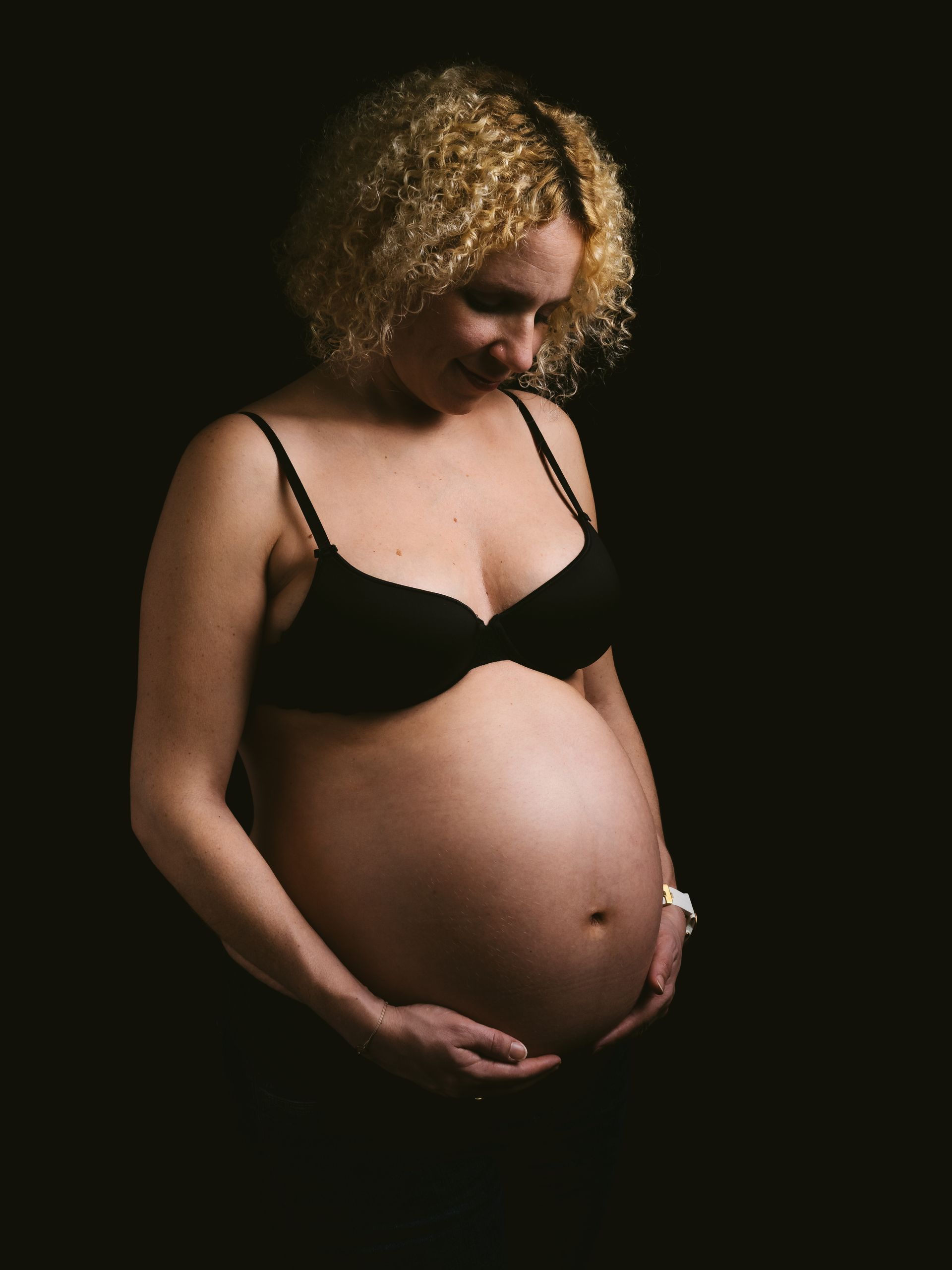 Schwangere Frau im schwarzen BH vor dunklem Hintergrund, die sanft ihren Bauch hält.