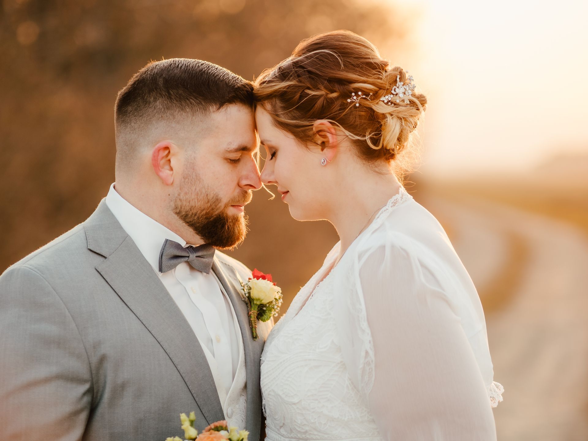 Ein Paar in Hochzeitskleidung berührt in einer sonnigen Außenkulisse die Stirn.