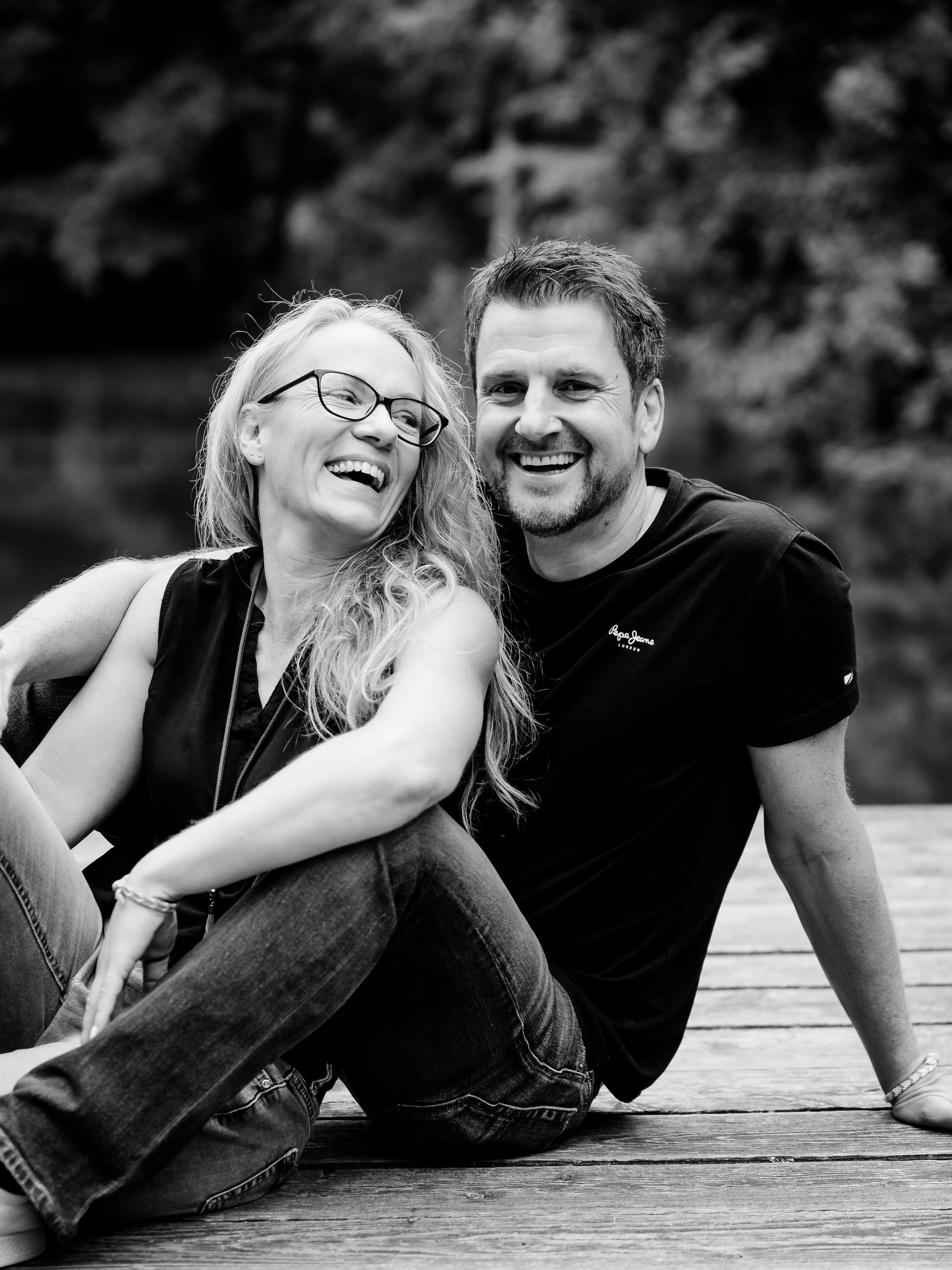 Zwei Personen sitzen im Freien eng beieinander, lächeln und lachen (Schwarzweißaufnahme).