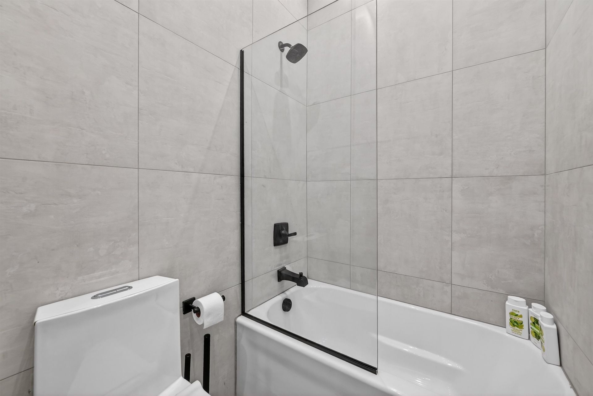 Modern bathroom with a toilet, bathtub, glass shower panel, and gray tiled walls