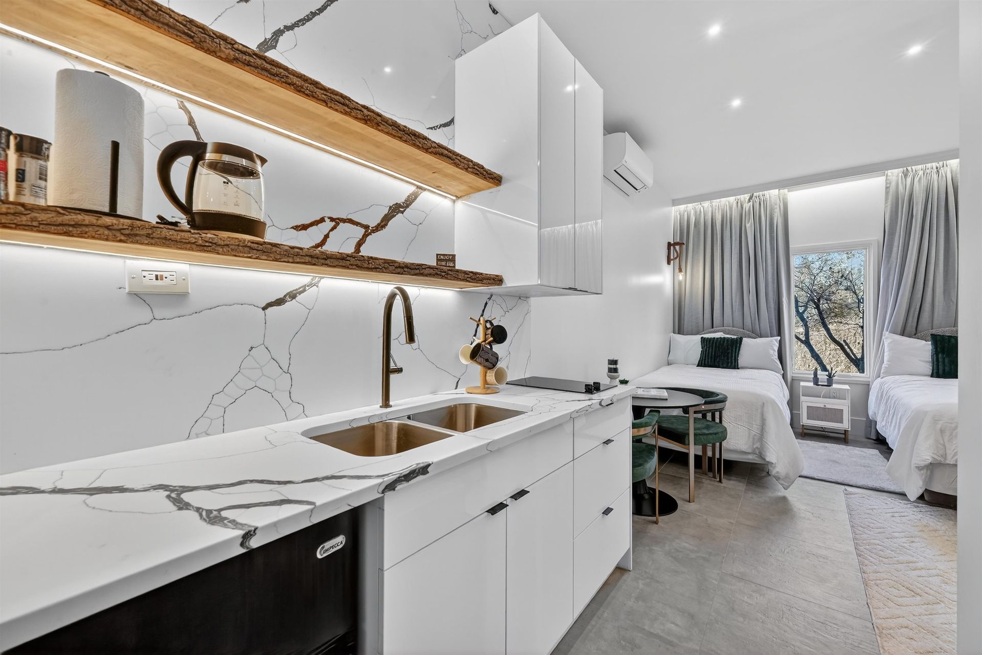 Modern white kitchen with marble counters, brass accents, and a bright living area beyond the window