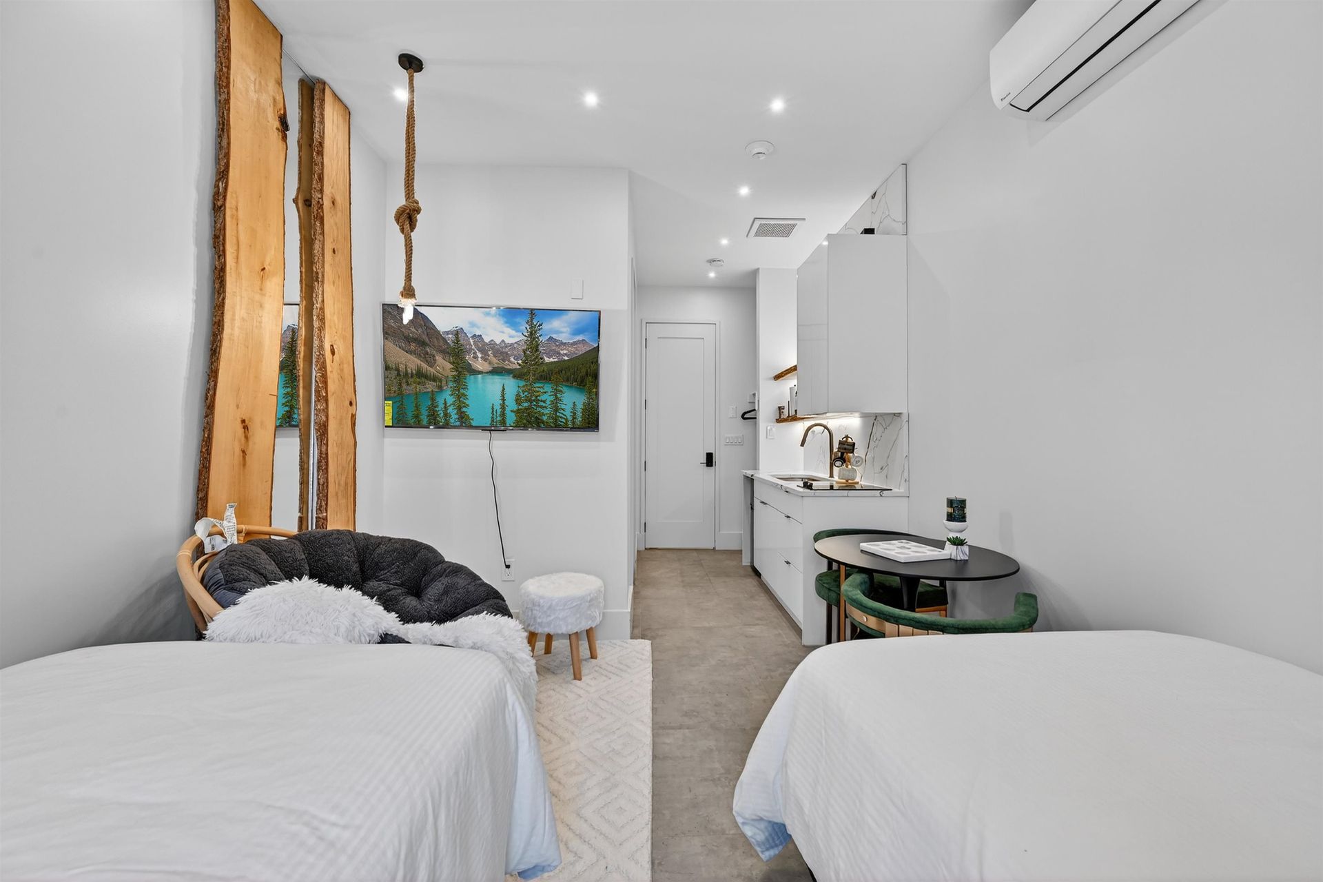 Bright bedroom with two beds, white walls, a wall-mounted TV, and a small kitchen area in the back.