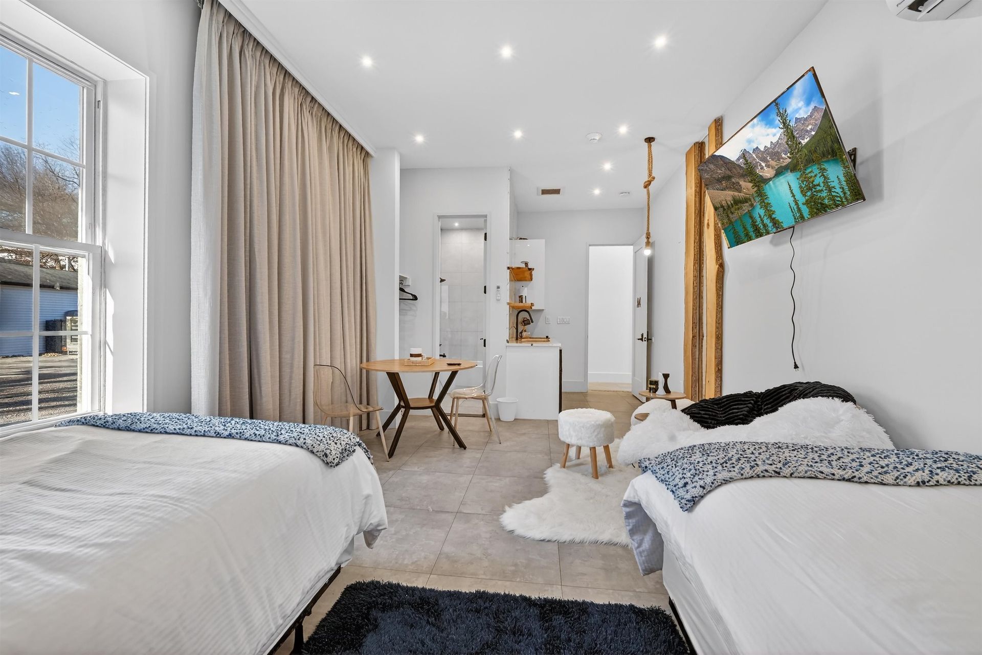 Bright modern bedroom with two twin beds, beige curtains, a TV, and a small desk near a window.