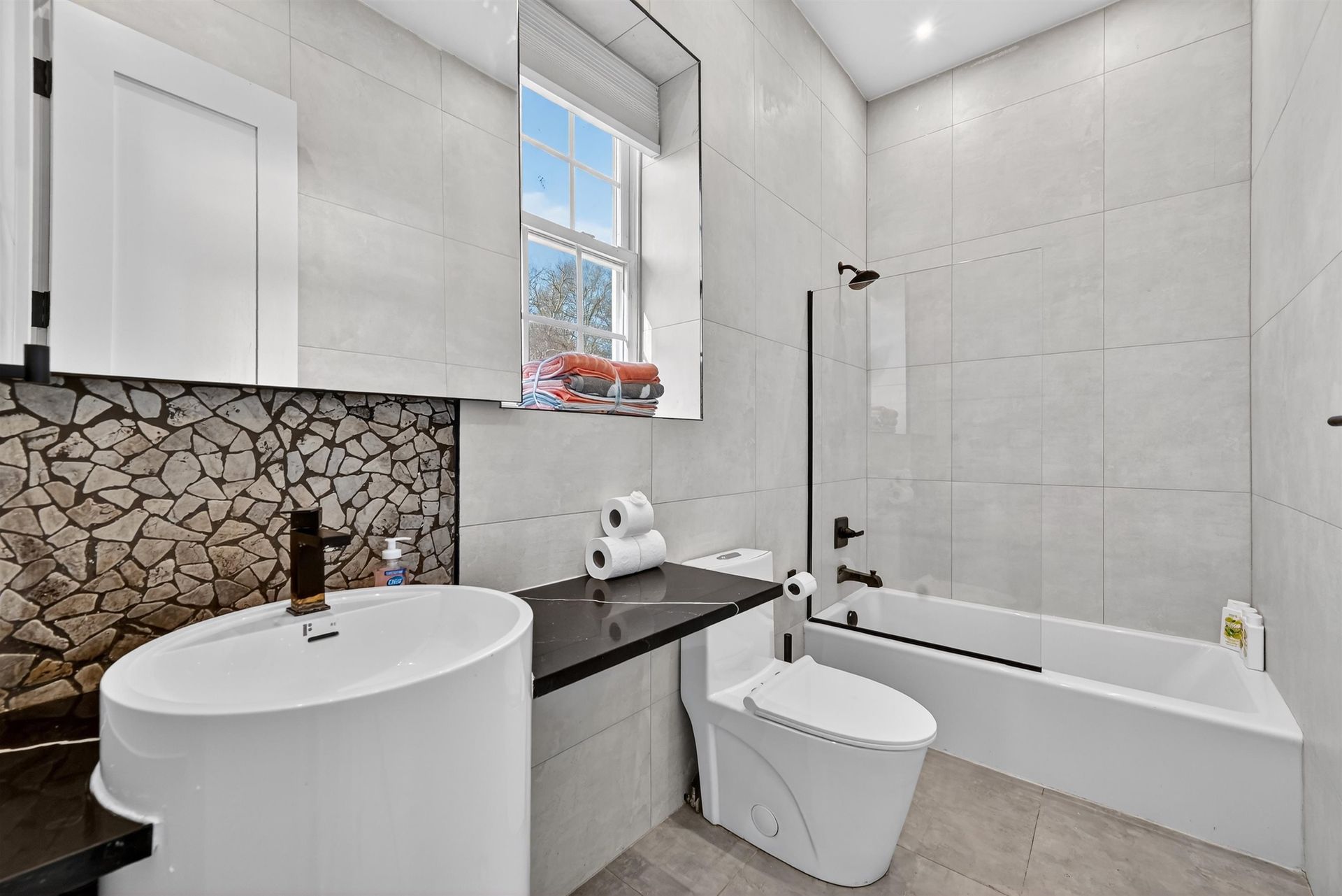 Modern bathroom with white tiles, freestanding tub, round sink, and window above the toilet