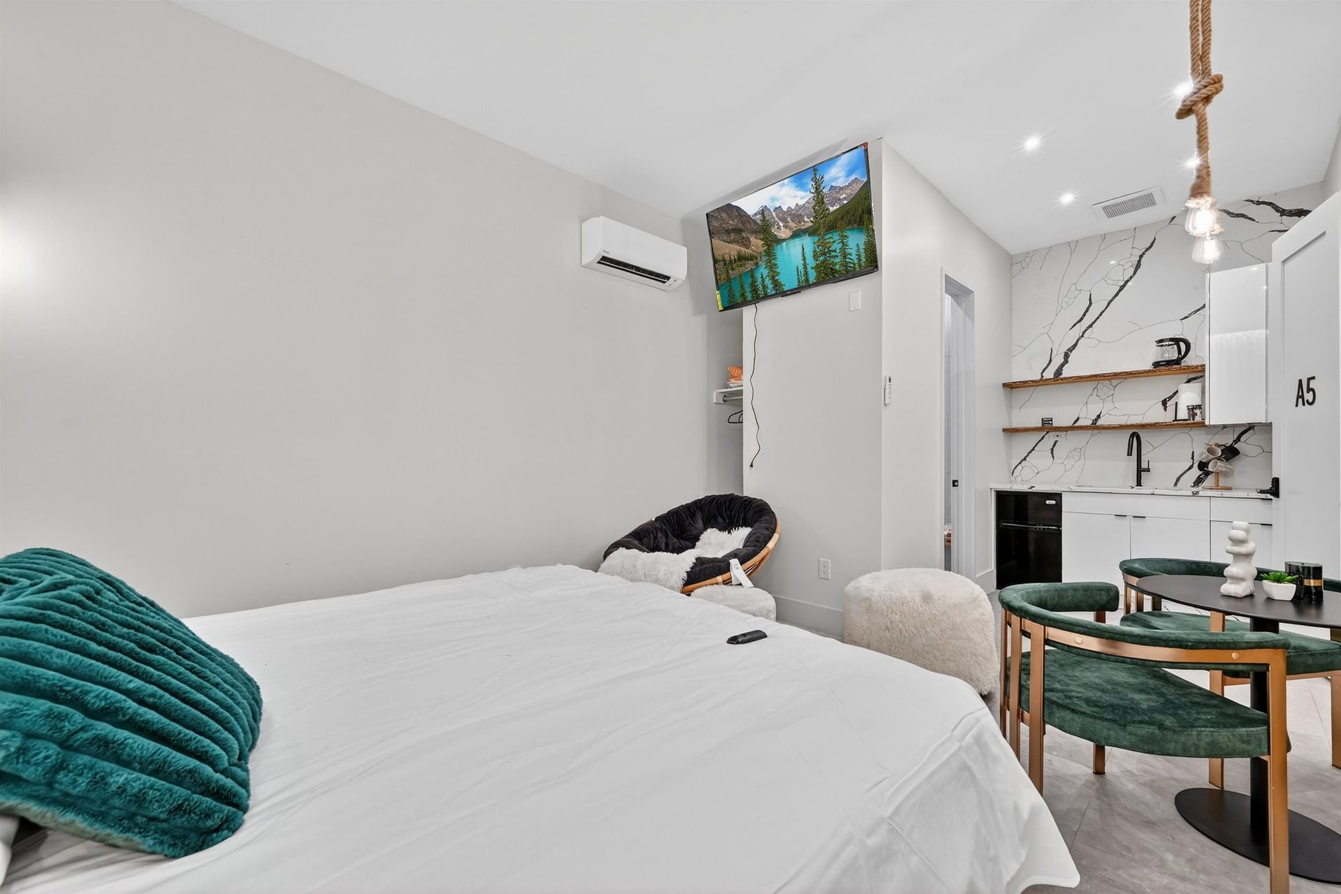 Bright minimalist bedroom with white bed, wall-mounted TV, fireplace, and a green-accented chair.
